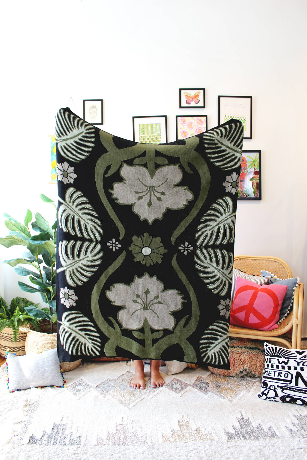 A knit blanket with a geometric pattern featuring floral-like designs in beige, black, and green, displayed over a wooden chair in a room with various decor.