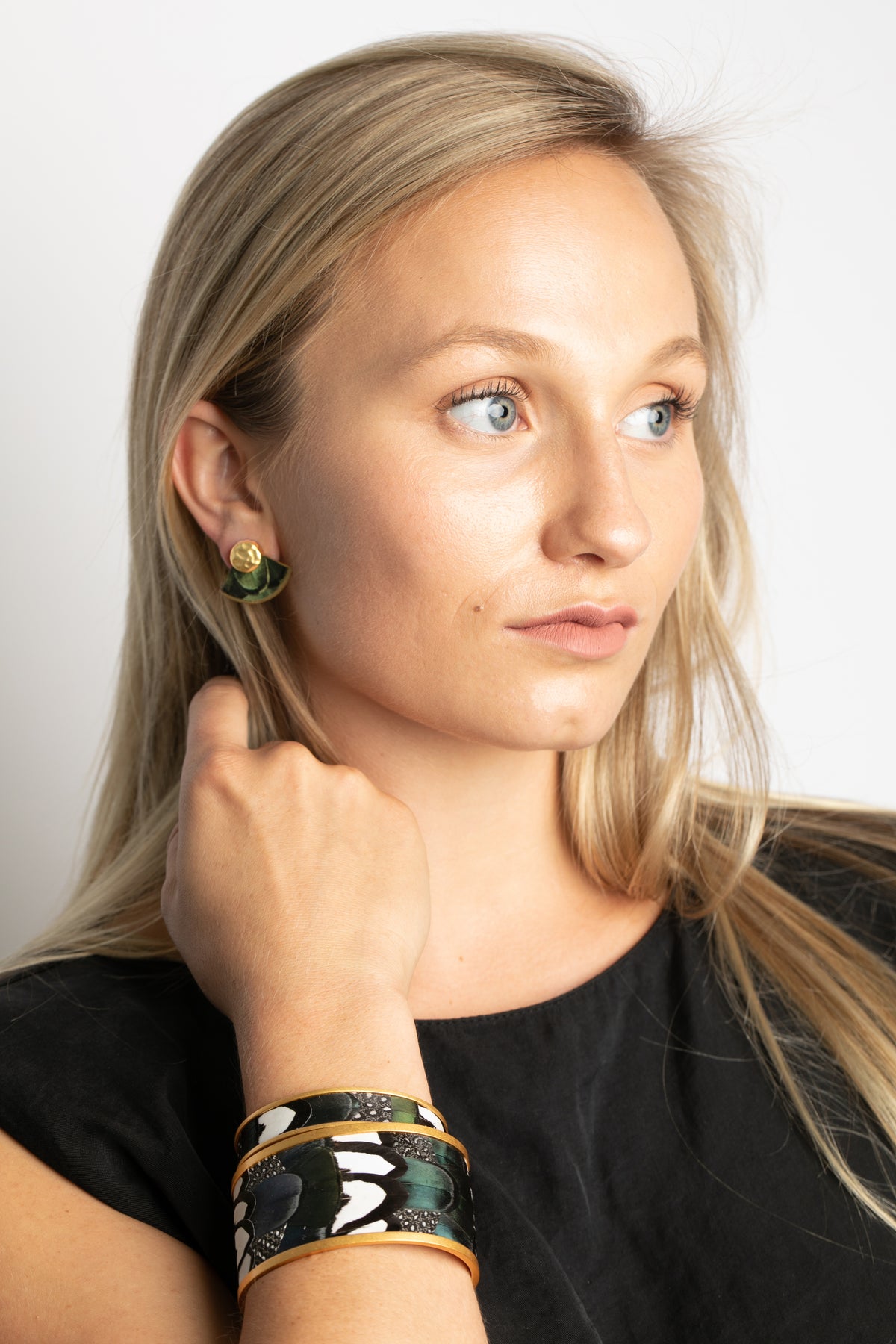 Woman wearing gold and feather earrings and bracelets on a plain background