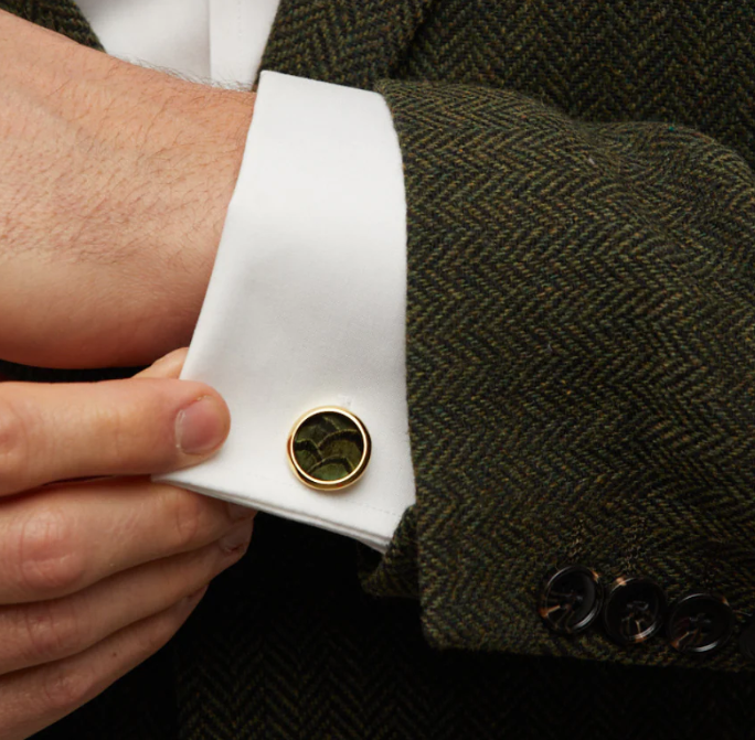 Close-up of a person wearing a round gold cufflink with green and black feather inlays on a white shirt with a green herringbone sports coat.