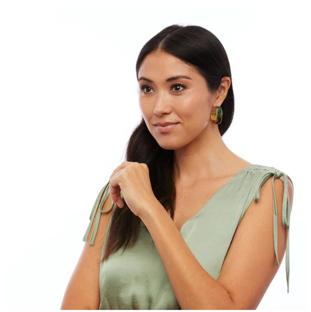 A woman with long dark hair wearing a sage green satin-like top with tie details on the shoulders, photographed against a plain white background. She is wearing elegant gold hoops with green and blue feather inlay