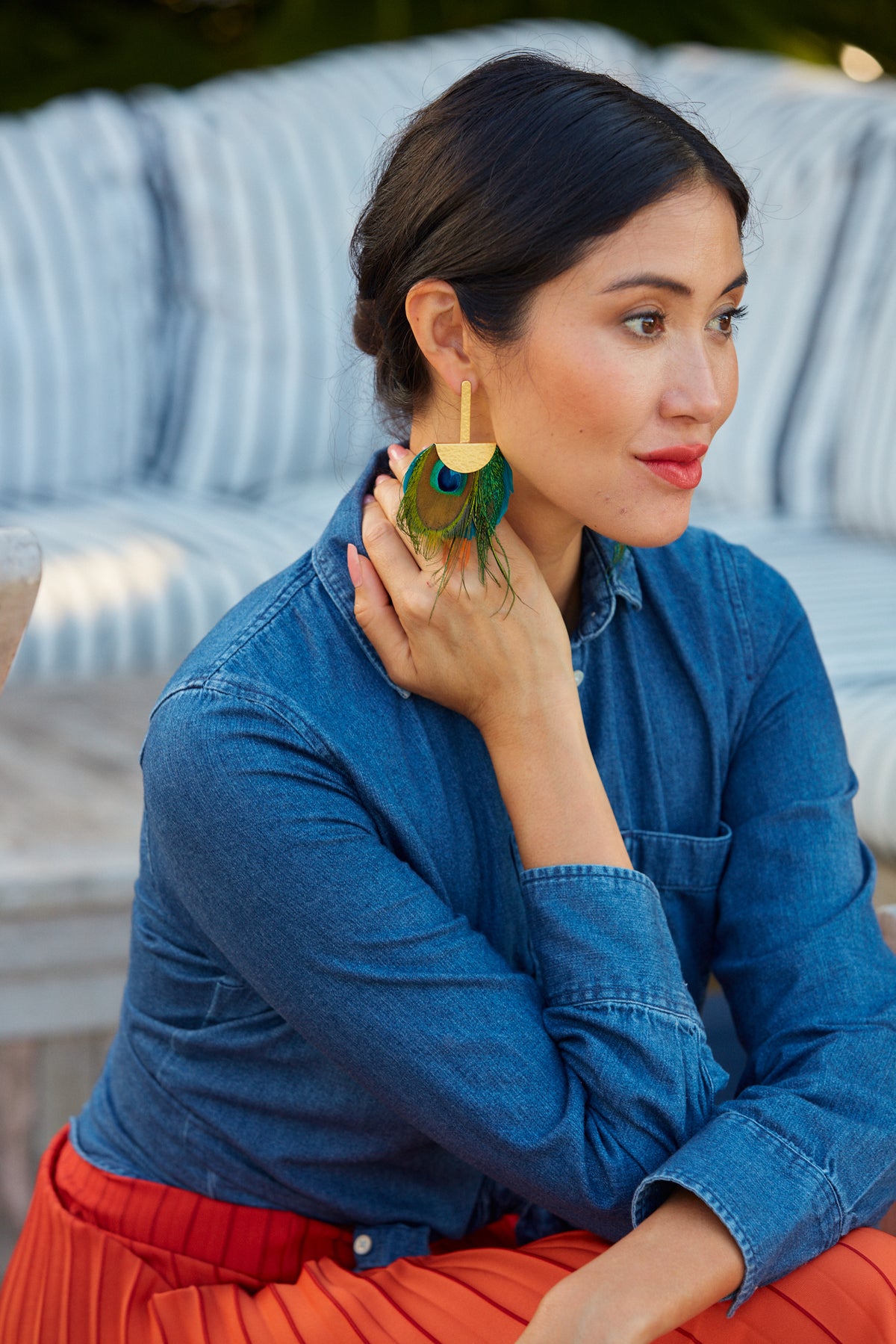 Woman in a blue denim top and red dress modeling a gold earring with dangling peacock feathers.