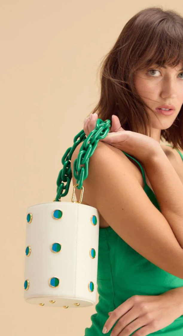 Woman in green dress holding a white leather bucket bag with chunky green chain hand strap. Bag features blue and green feather polka dot inlays around and gold foot hardware underneath