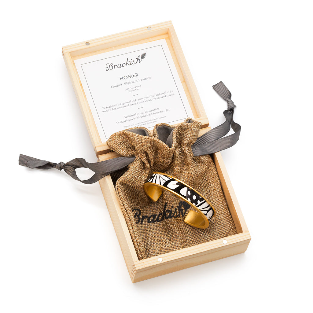 Gold cuff with black and white feather inlay in a wooden jewelry box containing an item description card and drawstring bag with Brackish branding.