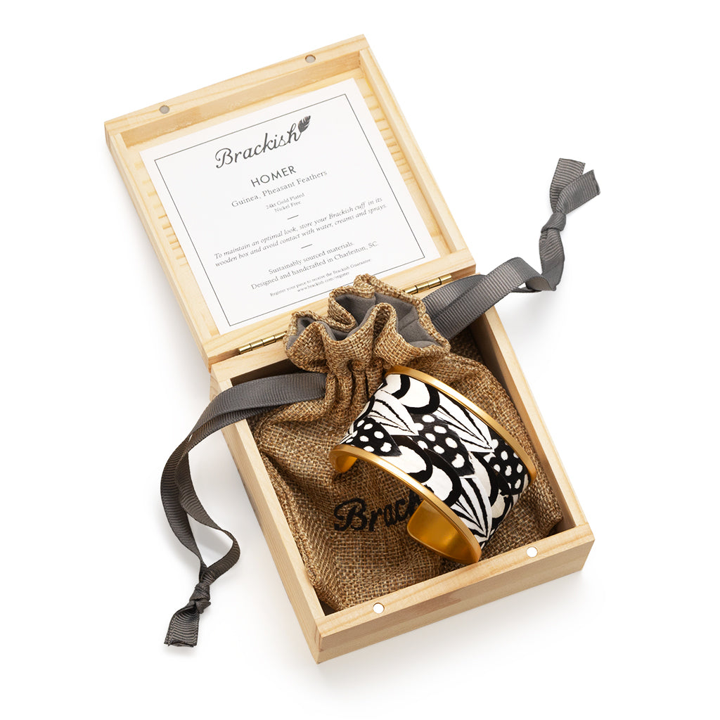 Wide gold cuff with black and white feather inlay shown in a wooden box with an item description card and brown drawstring bag with Brackish logo on a white background