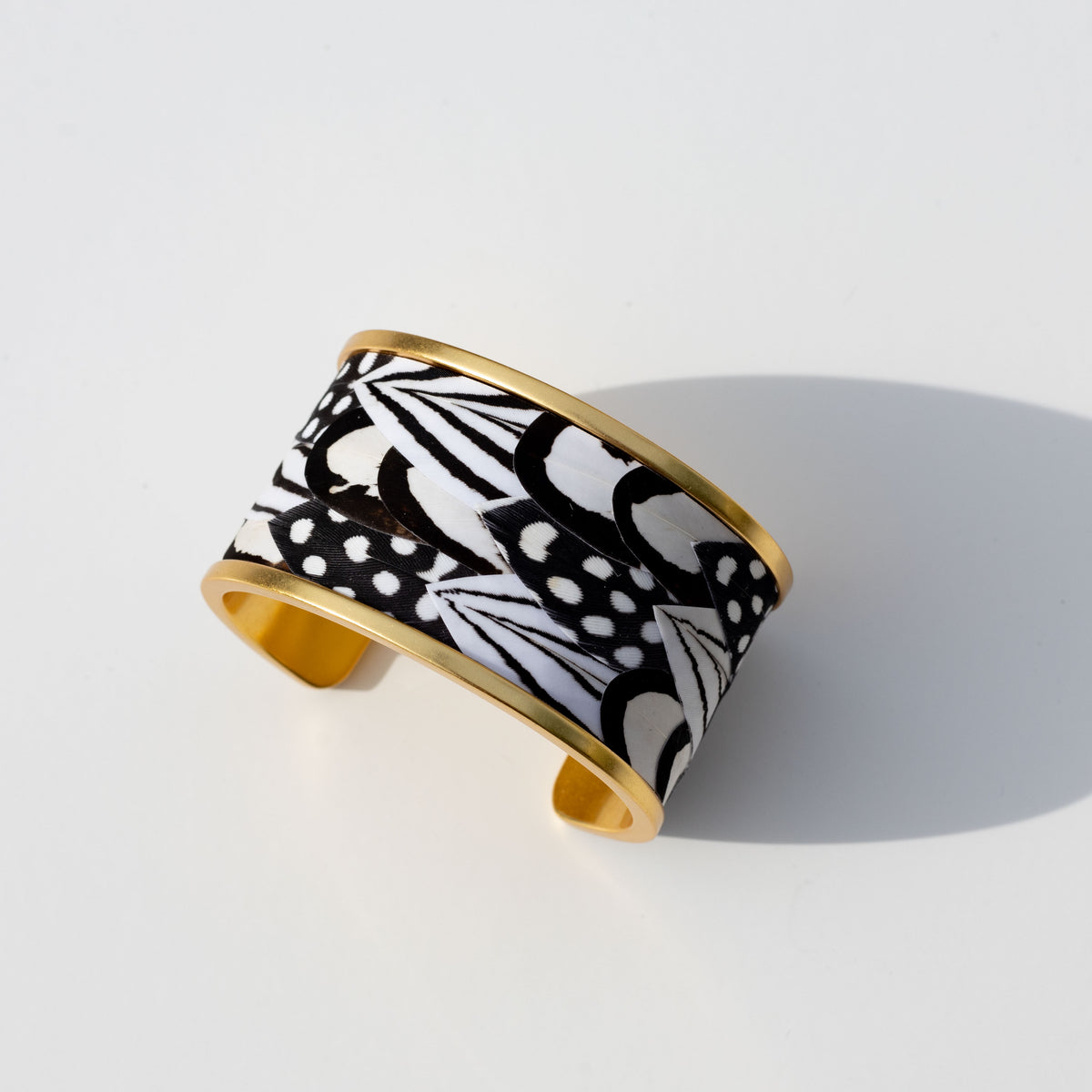 Overhead view of wide gold cuff with black and white feather inlay on a white background
