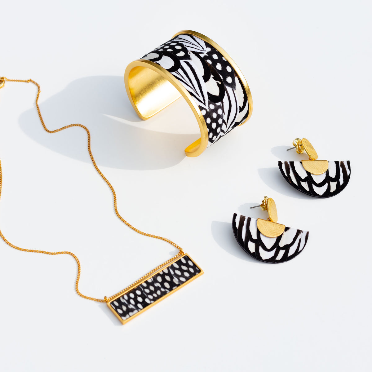 Matching set of gold jewelry, all with black and white feather inlays, showing a wide cuff, necklace, and earrings on a white background