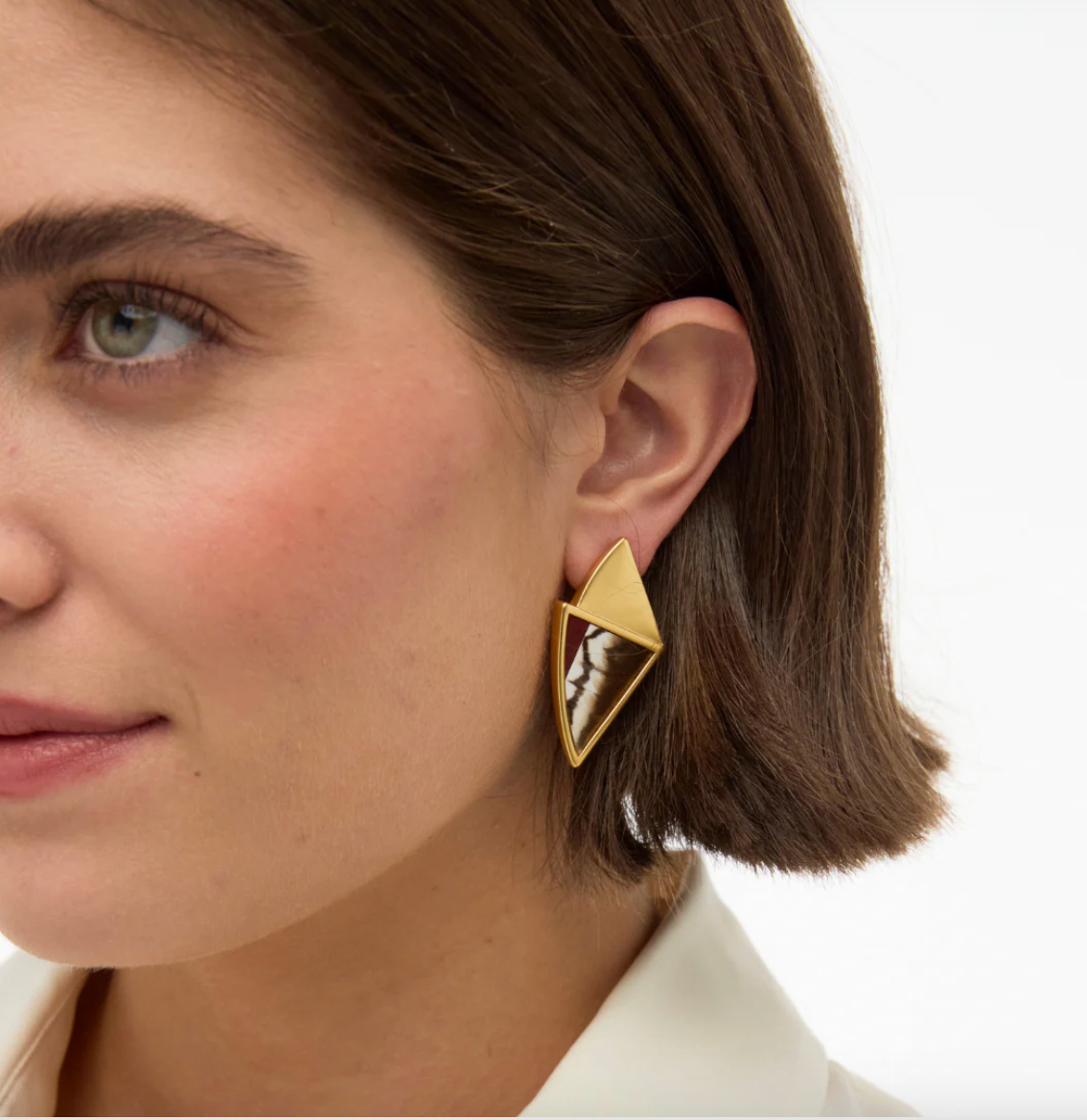 Close-up of a woman wearing diamond-shaped gold earrings with a red, brown and white feather inlay on a white background