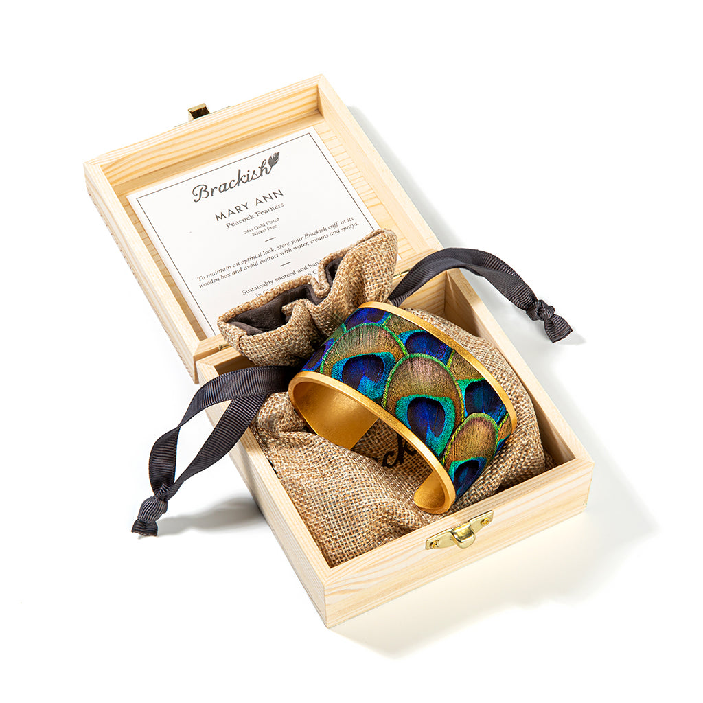 Wide gold cuff with peacock feather inlay shown in a wooden box with an item description card and brown drawstring bag with Brackish logo