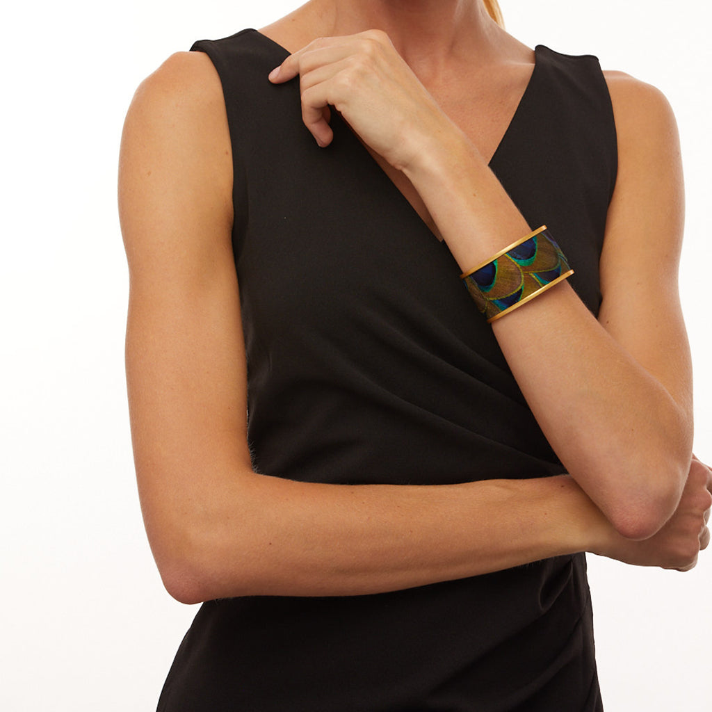 Woman in a black sleeveless dress modeling a wide gold cuff with peacock feather inlay against a white background