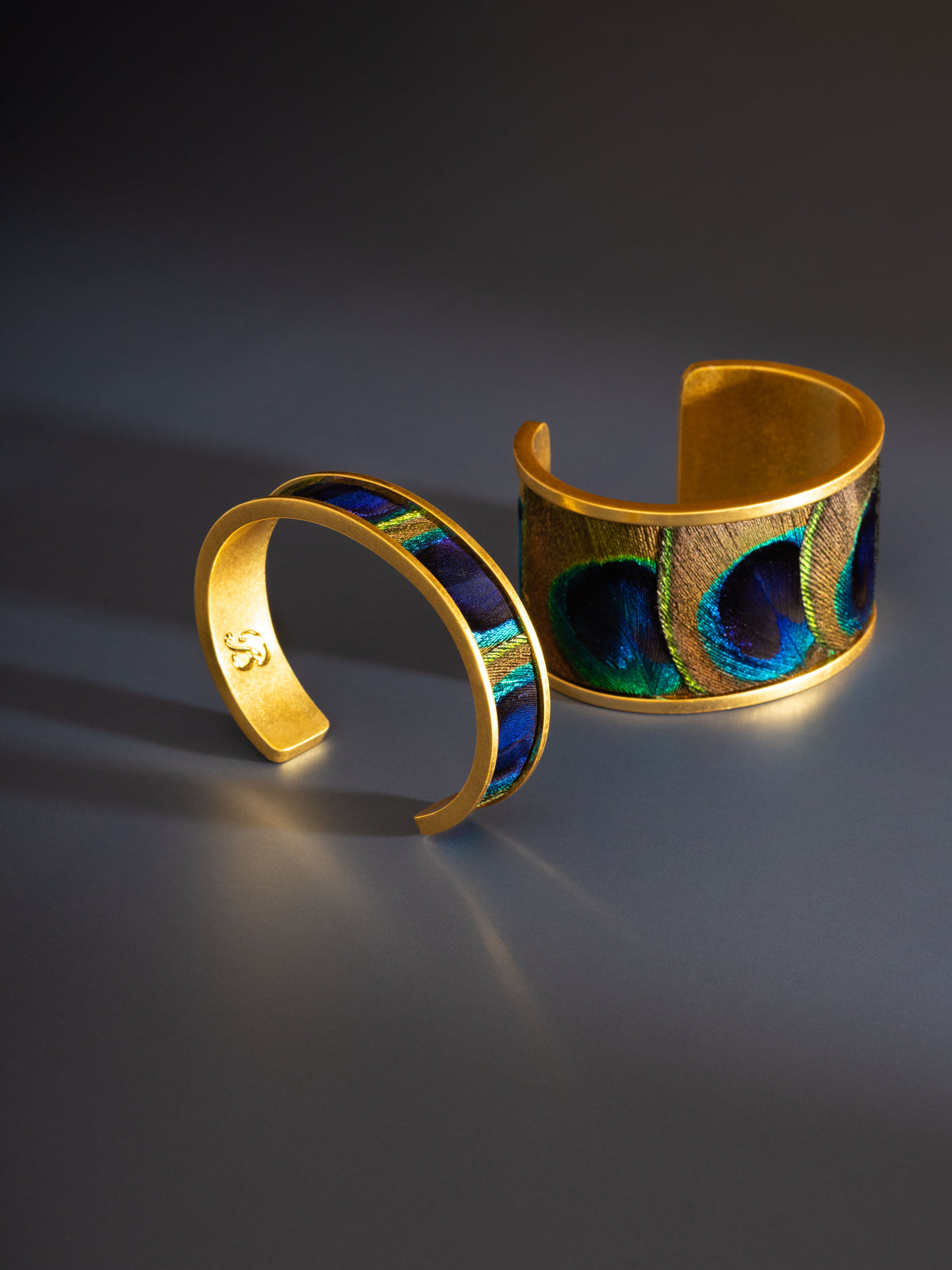 Two gold cuff bracelets, one thin, one wide, both with peacock feather inlay against a dark gray background