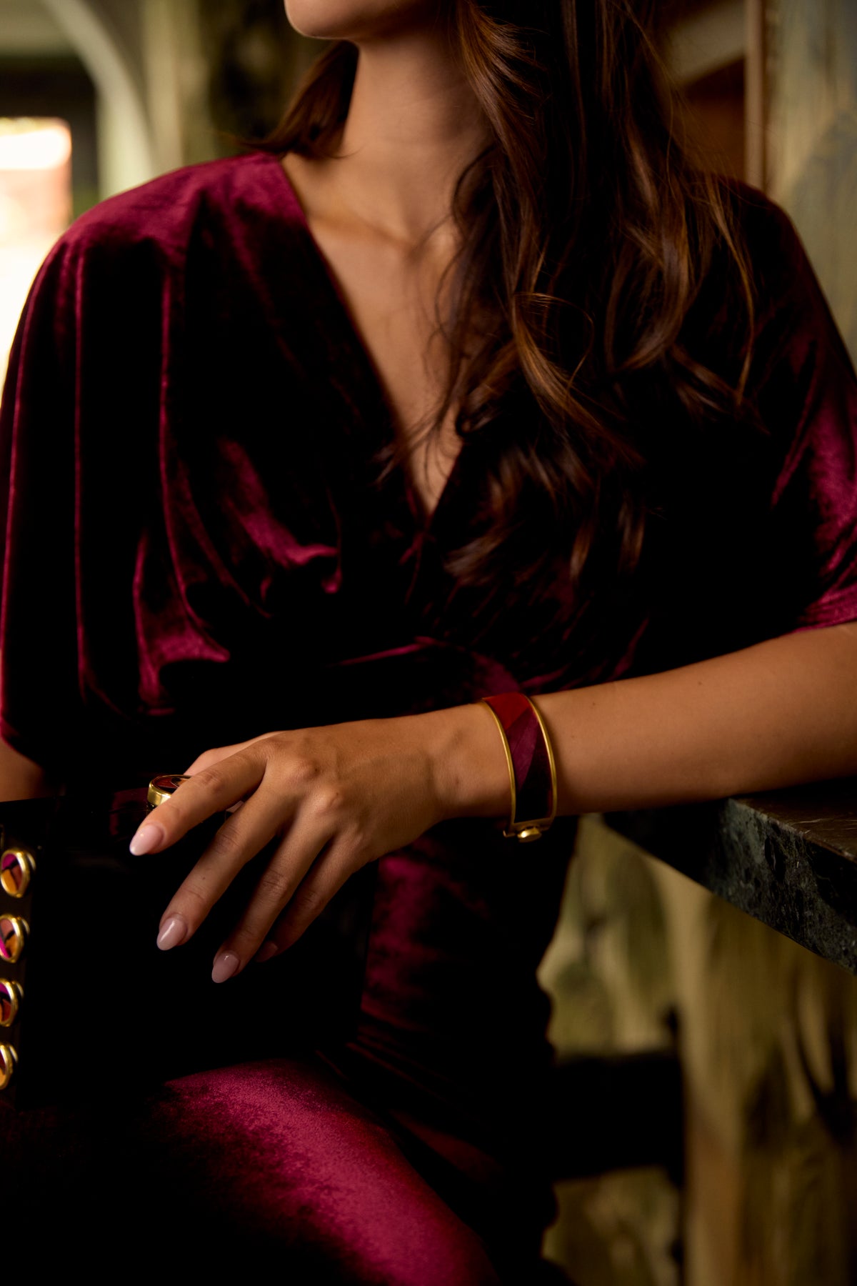 Woman in a dark red dress modeling a gold cuff with inlay of red and purple feathers.