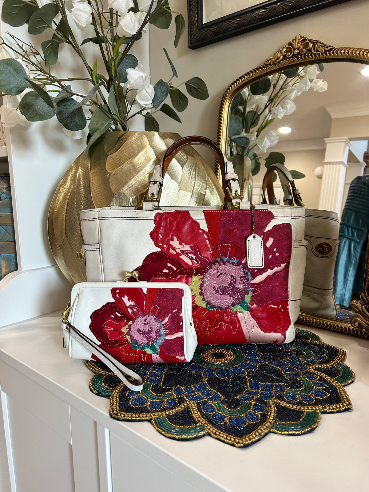 A white handbag and matching wristlet clutch with a bold red and pink floral print, styled on a white console table with a decorative rug, gold mirror, and artificial flowers.