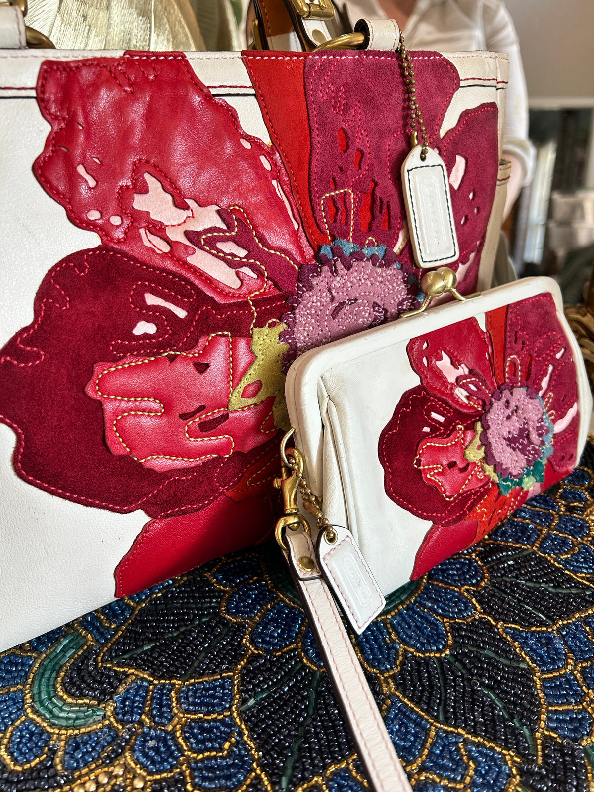 A close-up of a handbag and matching wallet featuring large floral applique in red, pink, and burgundy on a white base, with gold-tone hardware and a hanging tag.