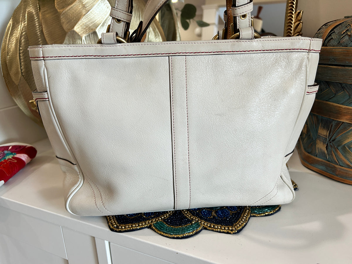 A large off-white leather handbag with double vertical seams and gold-tone hardware, sitting on a white surface with decorative items in the background.