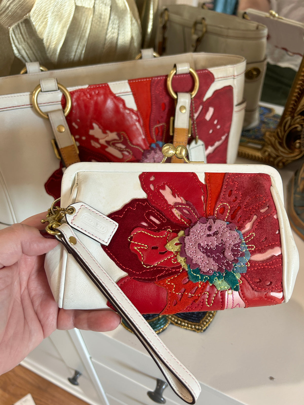 A hand holds a small white and red floral patchwork wristlet clutch with a white leather wrist strap and gold-tone hardware, with a larger matching patchwork tote bag in the background.