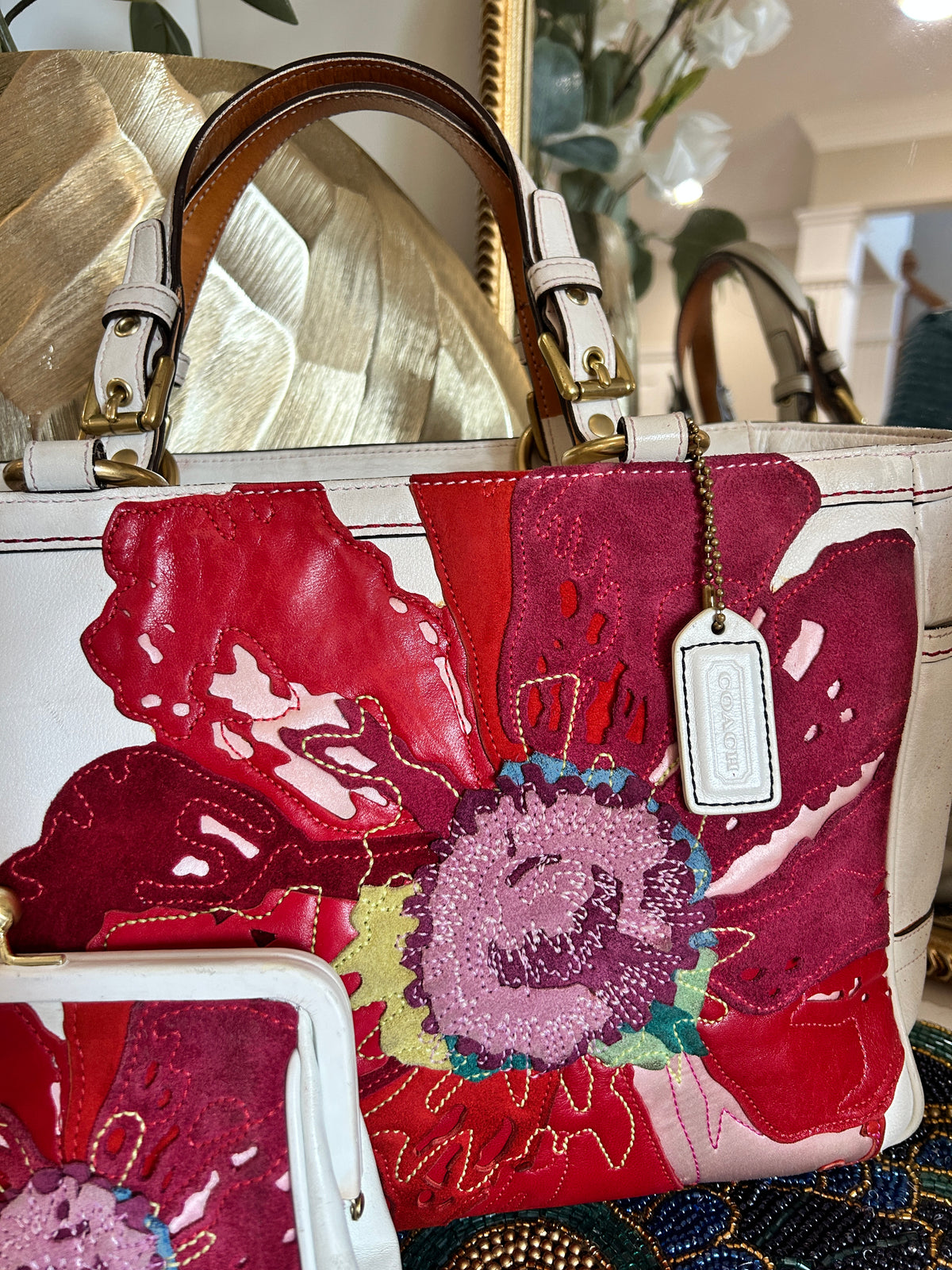 A white leather handbag with large, vibrant red and pink floral applique patches on the front, featuring gold-tone hardware, a hanging brand tag, and a matching coin purse partially visible in the lower left corner.