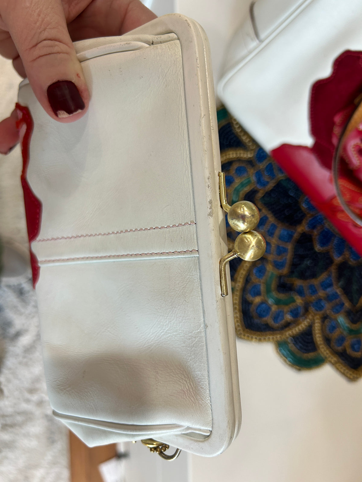 A close-up of a hand holding a small white leather kiss-lock clutch purse with gold-tone metal clasps. The hand has dark red nail polish, and other colorful items are partially visible in the background.