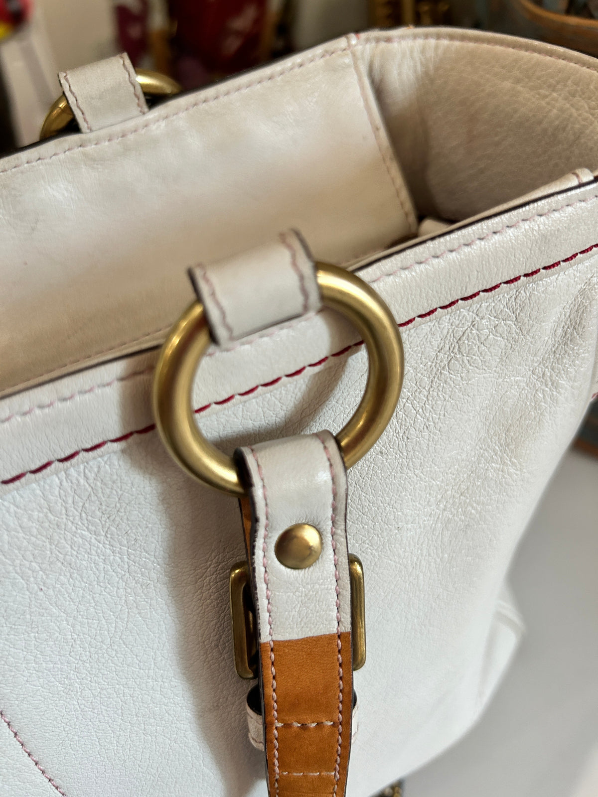 Close-up of a white leather handbag with a large gold-tone O-ring handle, contrast red stitching, and a tan leather strap detail.