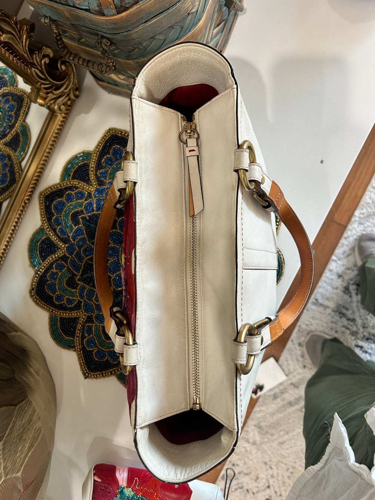 A top-down view of a cream-colored leather handbag with tan leather handles and gold-tone hardware, displayed on a white surface next to decorative items.