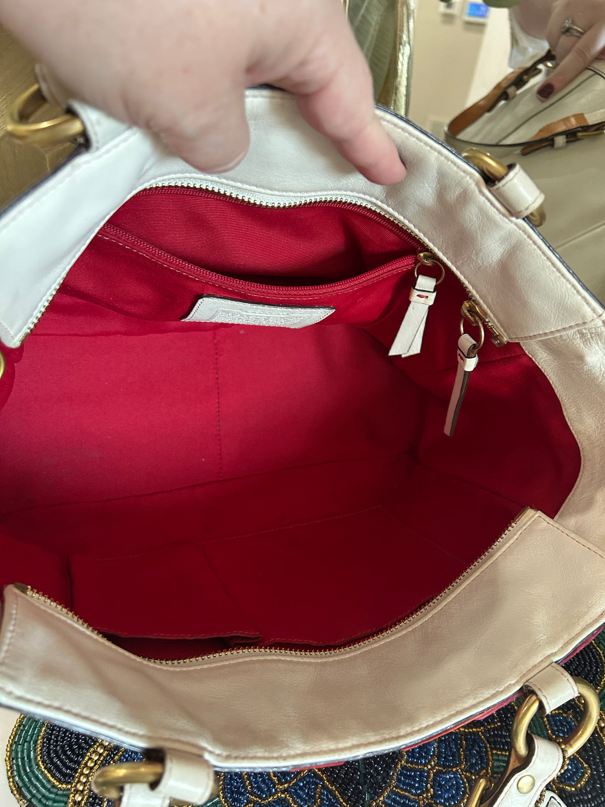 Close-up of the inside of a handbag being held open by a hand. The bag has a cream-colored exterior, a bright red interior lining, and gold-tone hardware.