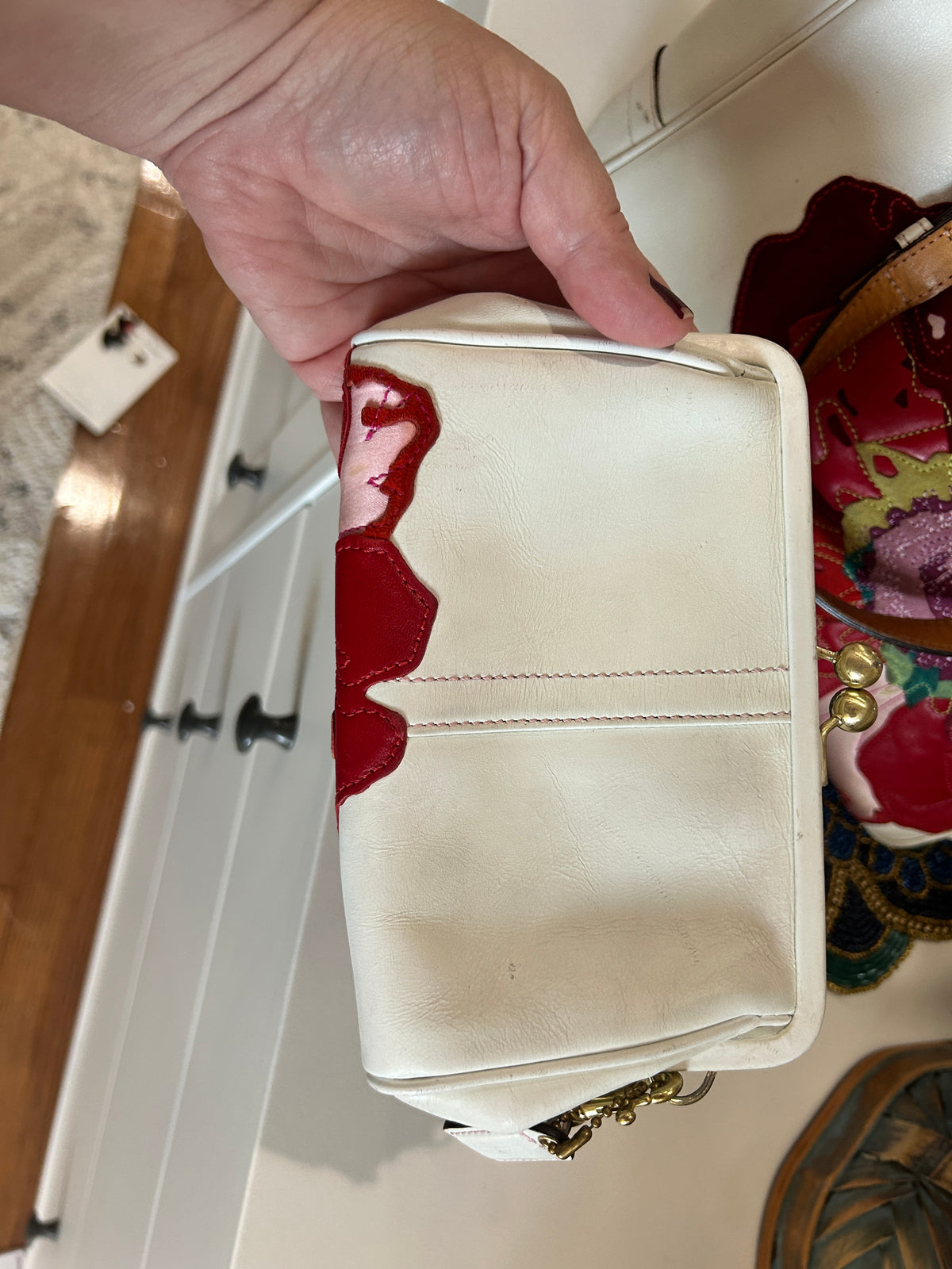 A hand holding a cream-colored leather clutch bag, set against a domestic background with white cabinets.