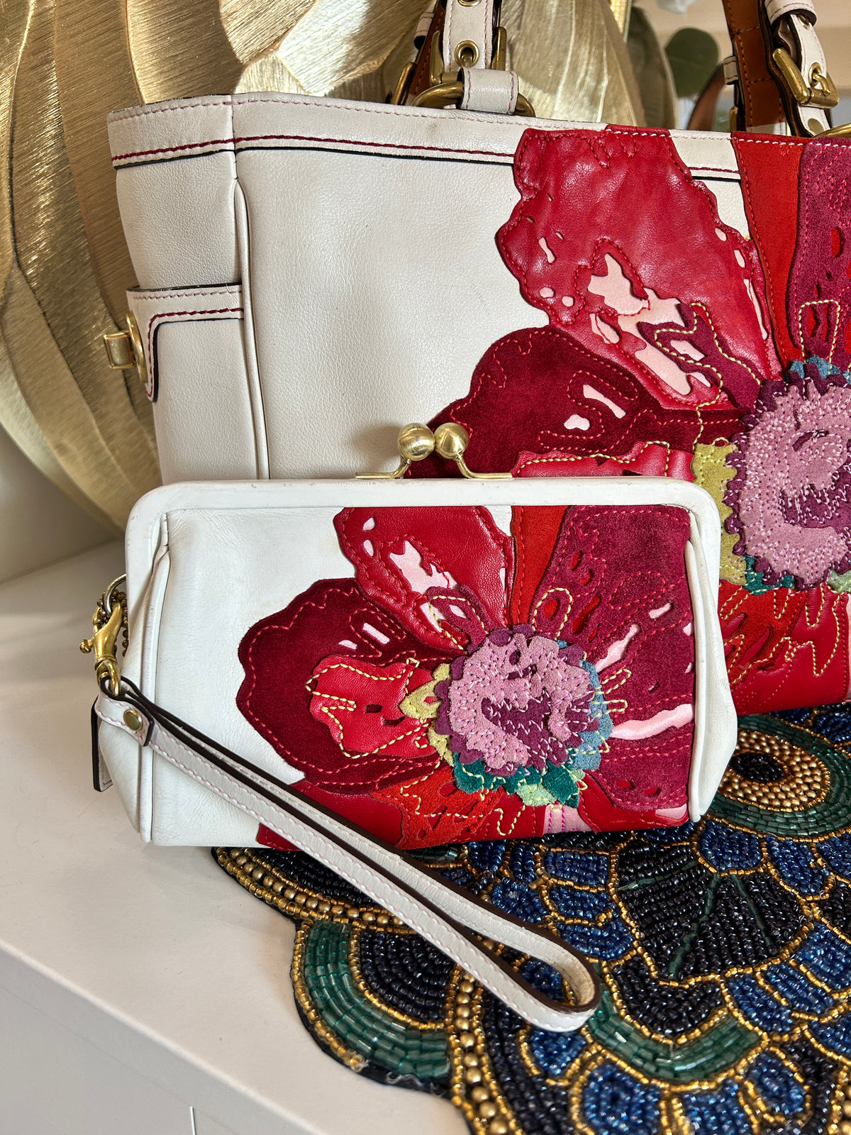 A white leather wristlet clutch with a matching wrist strap, decorated with a large red and pink floral patchwork applique, displayed in front of a larger matching handbag.