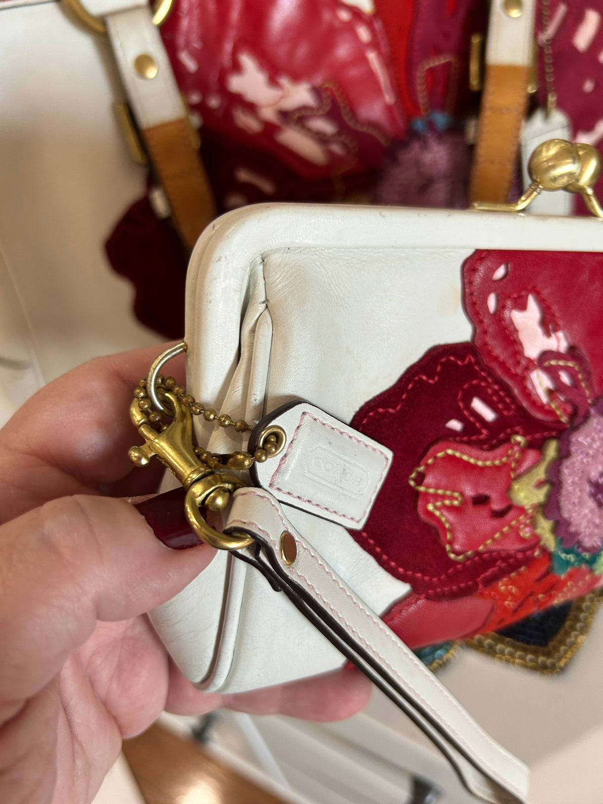 A close-up of a hand holding a white leather kiss-lock wallet with gold-tone hardware and a red floral patchwork detail on the front.