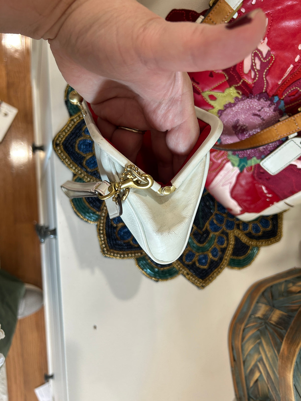 A close-up of a hand holding the opening of a small cream-colored leather clutch bag with gold-tone hardware, set against a background of other colorful bags on a white surface.