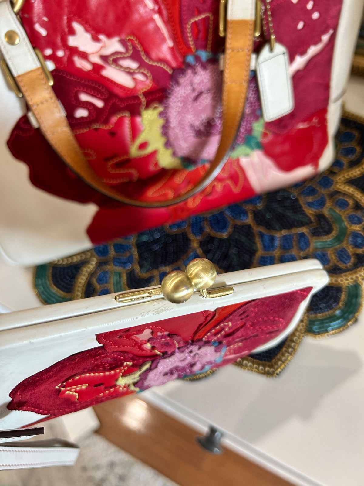 A close-up of a white clutch bag with a gold kisslock clasp and a red floral embroidered design, with another red and white handbag partially visible in the background.