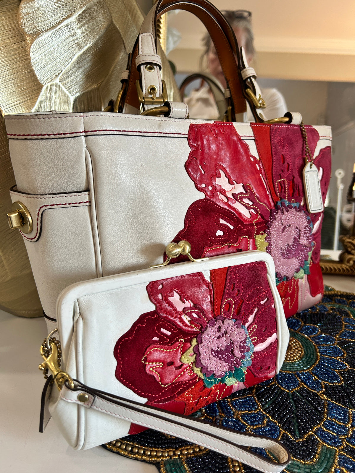 A white leather handbag and a matching kisslock wristlet, both decorated with large red and pink floral patches, shown in a styled indoor setting.