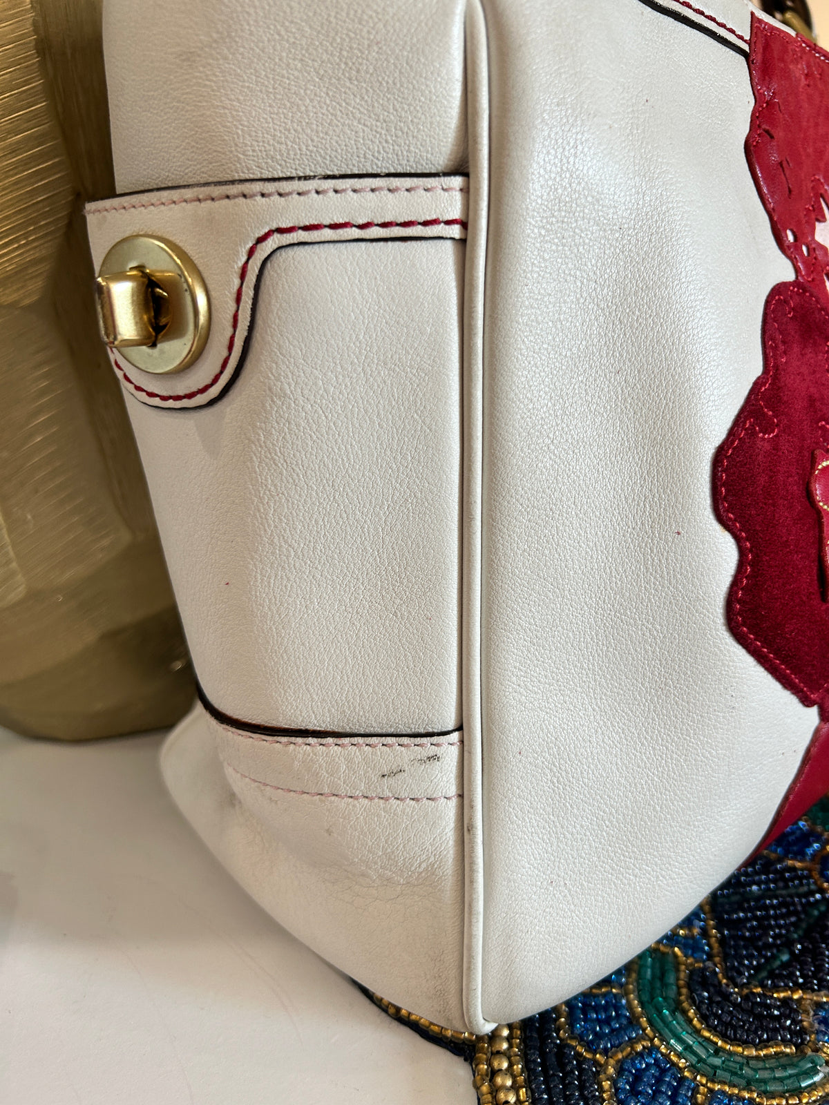 Close-up of a white leather handbag showing a side pocket with red contrast stitching and a gold turnlock closure.