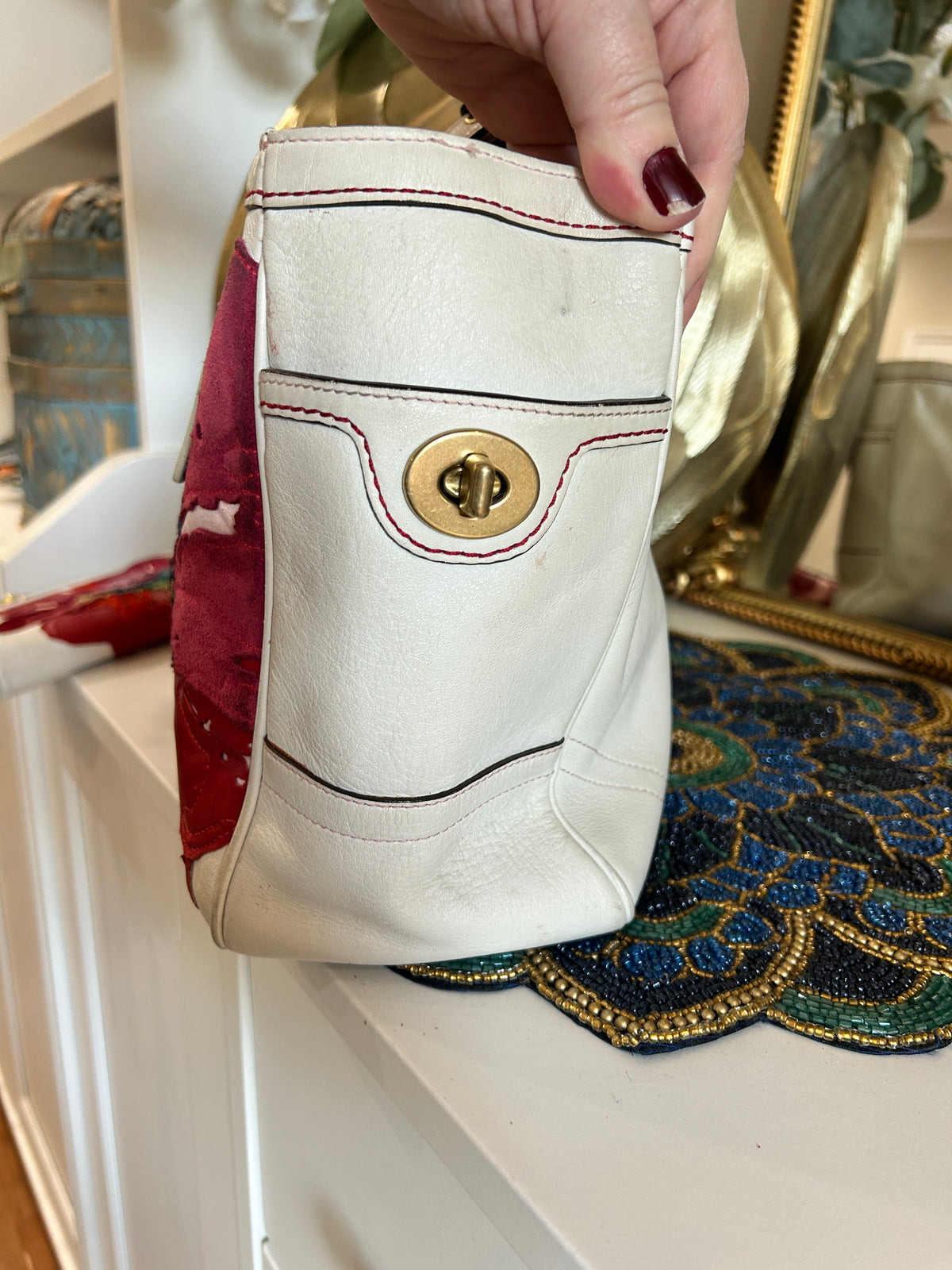A hand with dark red nail polish holds a small white leather pouch with a gold-tone turn-lock closure and decorative red and black stitching. The pouch is positioned over a white cabinet, with a colorful patterned textile and a gold-framed mirror in the background.