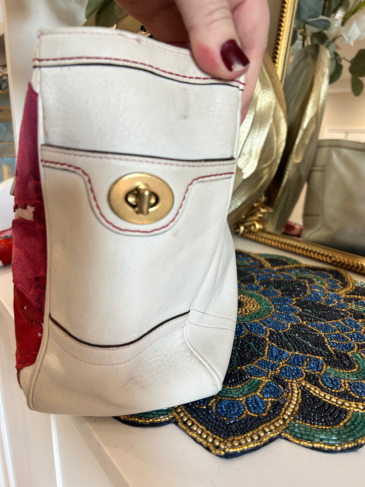 A close-up of a white leather handbag with a gold-tone turn-lock clasp and red/black contrast stitching, held by a hand with dark burgundy nail polish over a decorative blue and gold beaded surface.