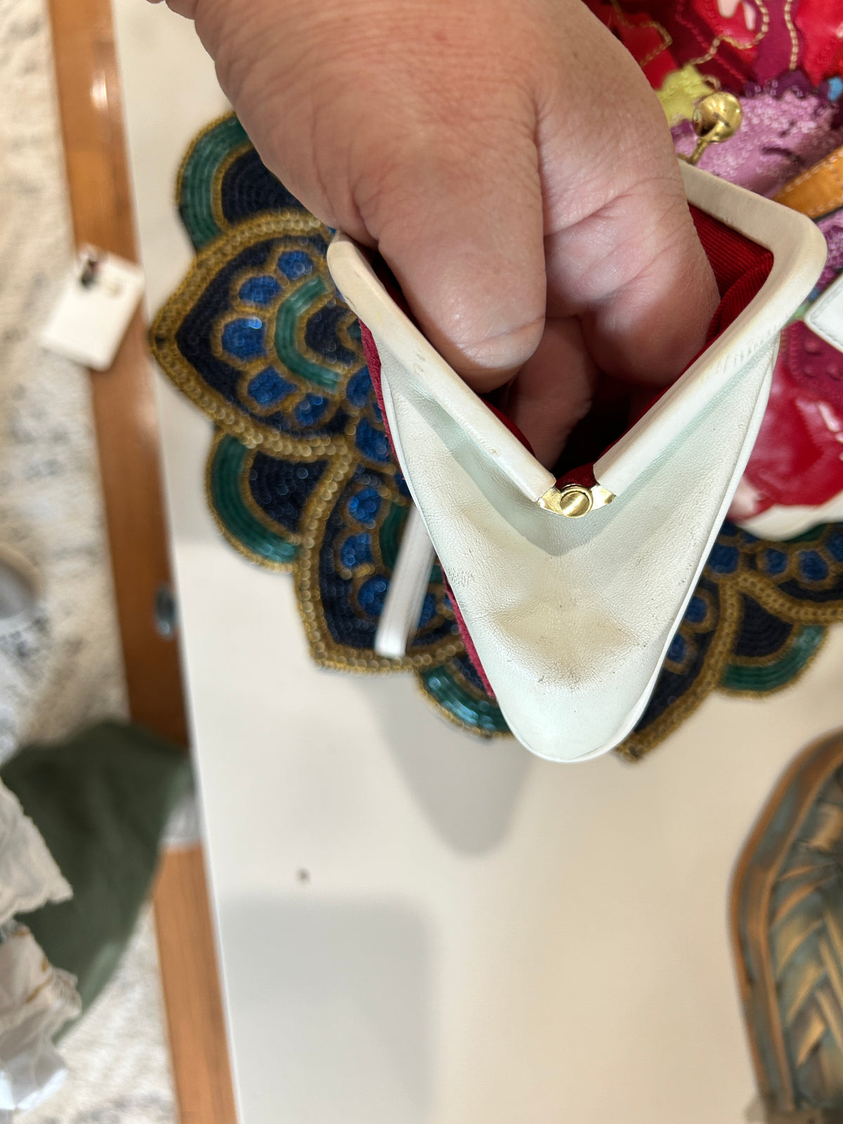 A close-up of a hand holding a cream-colored, triangular vintage clutch purse with a gold-tone clasp. The purse is positioned in front of a colorful embroidered textile.
