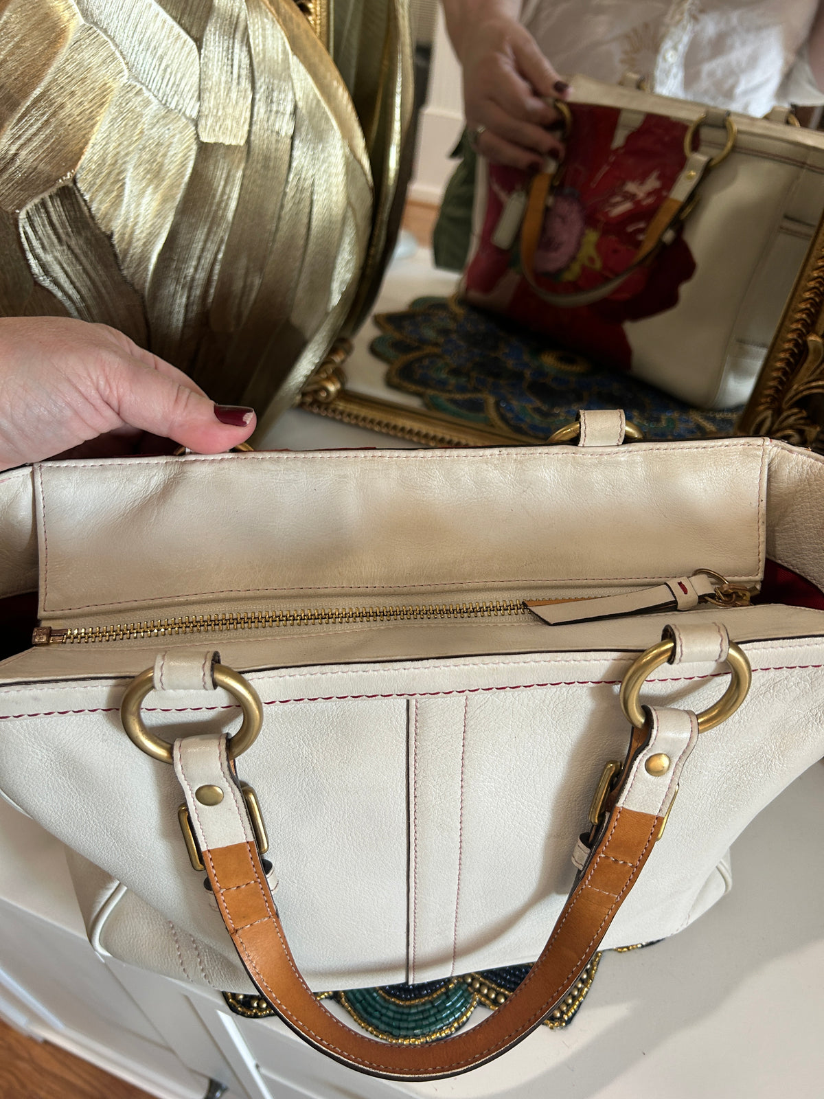 A cream-colored leather handbag with tan leather handles and gold-tone hardware, shown with a hand holding it open in front of a mirror reflecting another view of the bag.