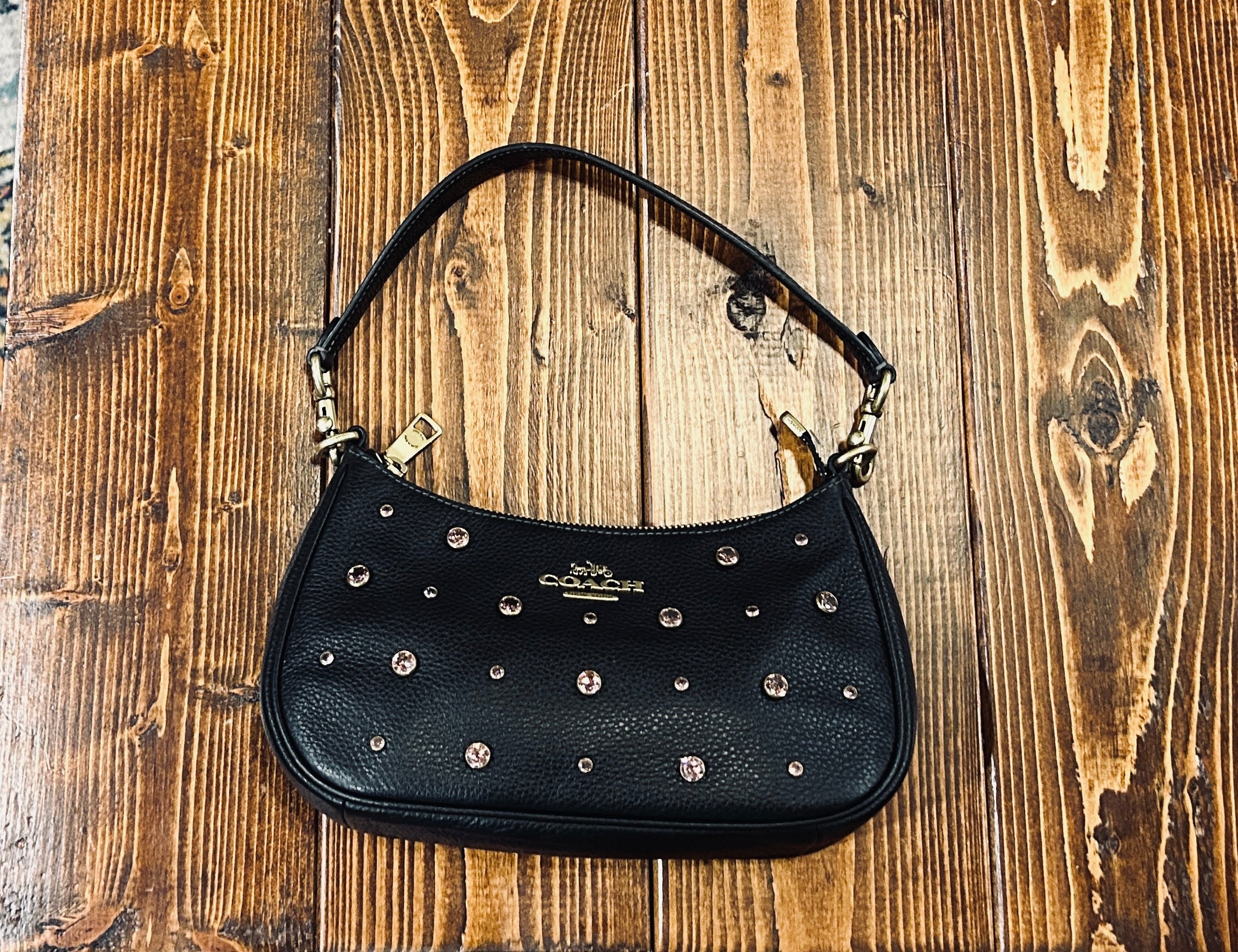 A black pebbled leather Coach shoulder bag with pink faceted crystal studs and a gold-tone logo on the front, displayed on a rustic wooden plank surface.