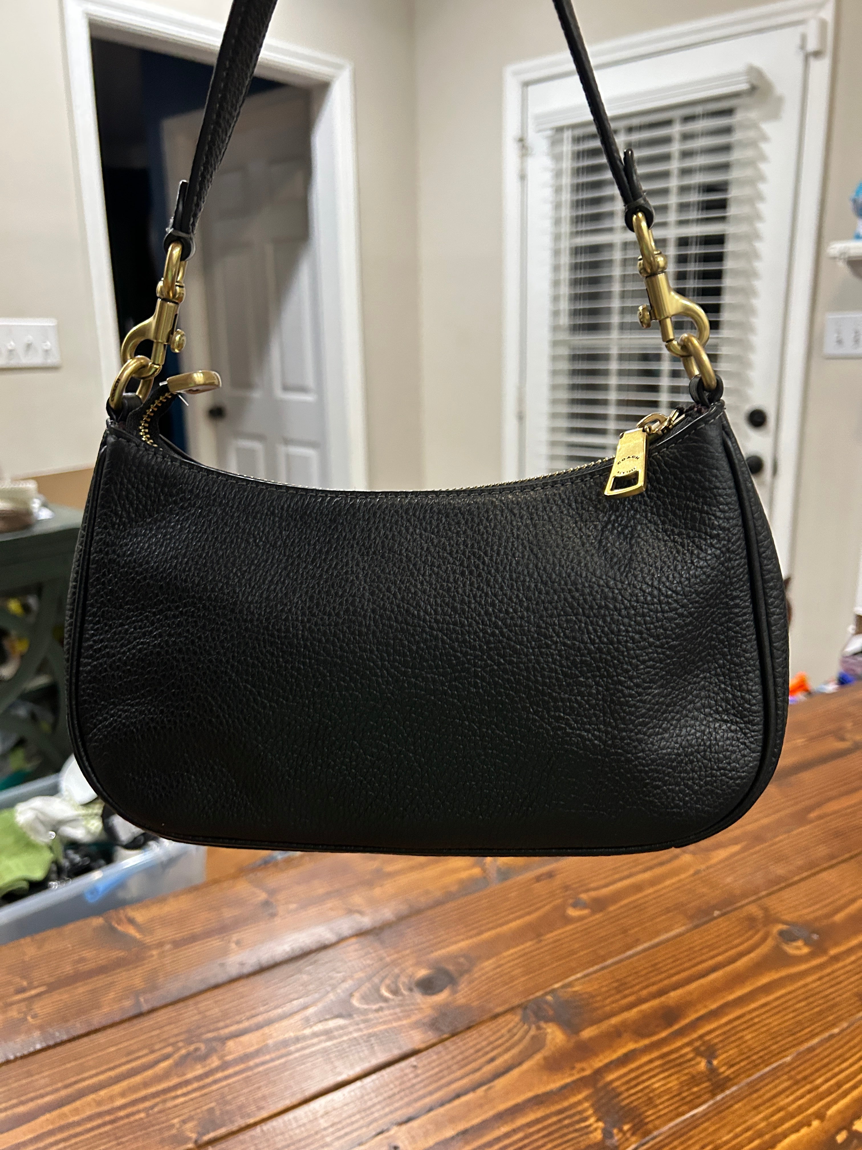 A black pebbled leather shoulder bag with gold-tone hardware, hanging in front of a wooden table in a home interior.