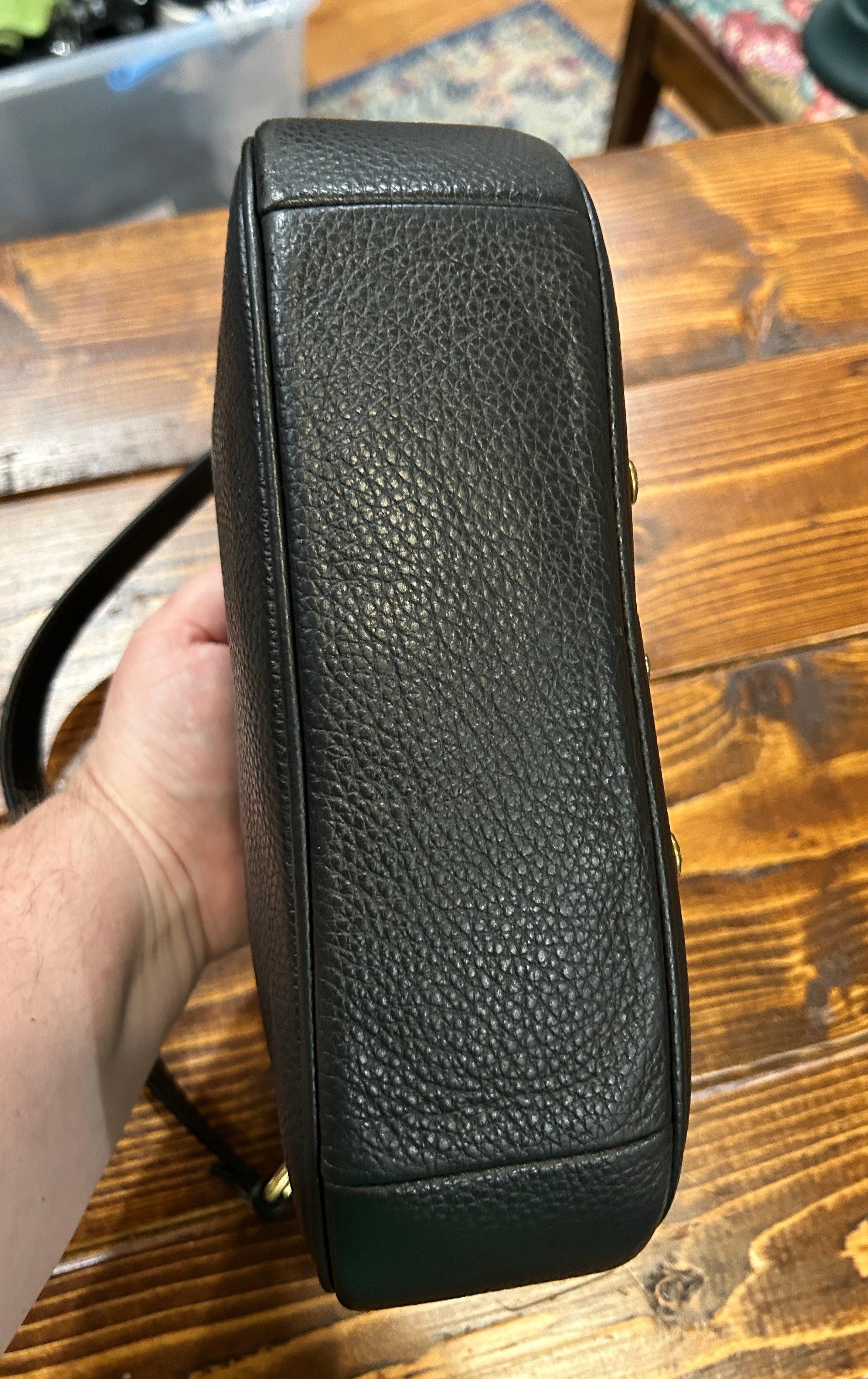 A close-up of a black pebbled leather handbag being held by a hand over a wooden table.