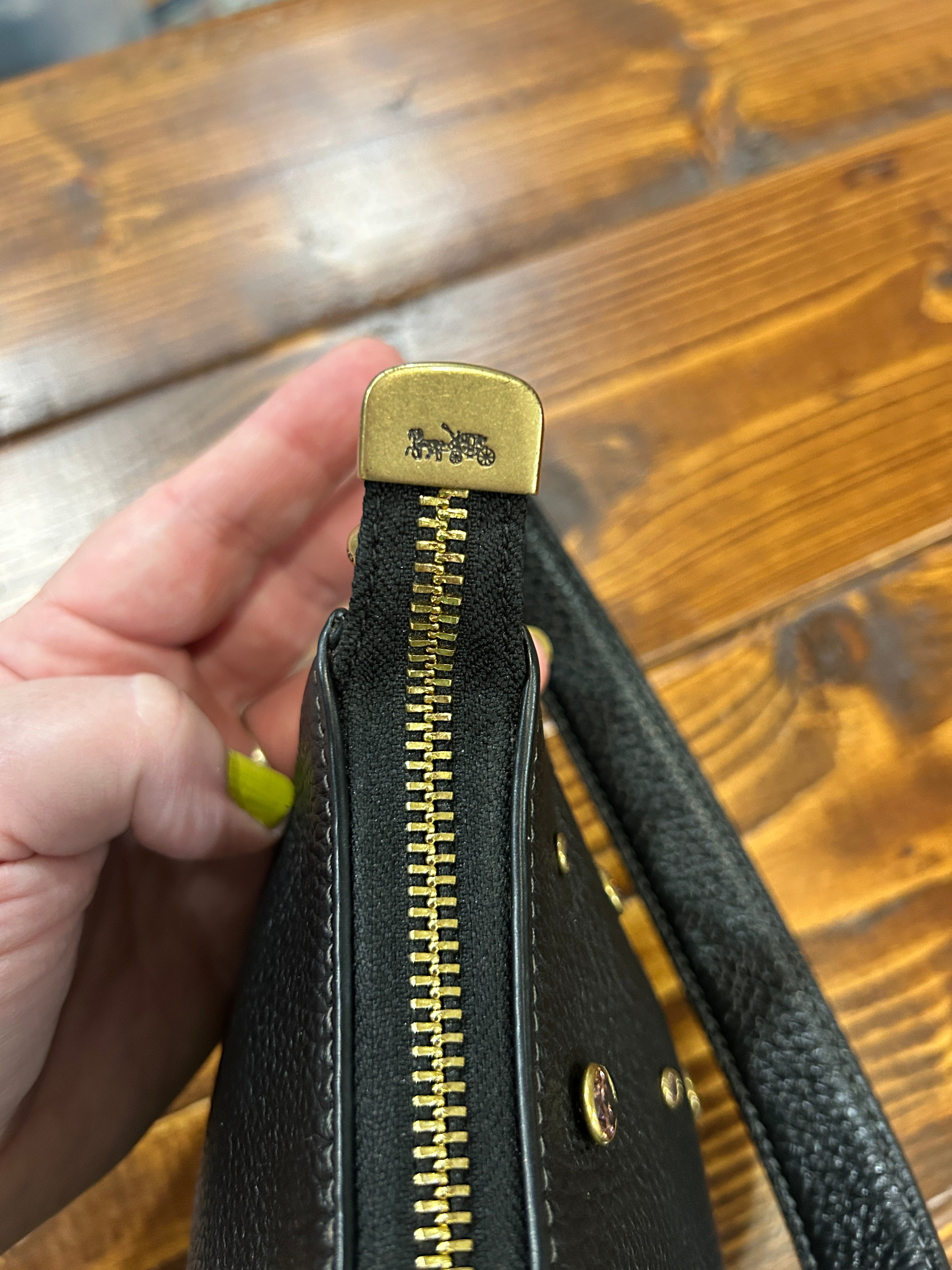 A close-up of a black pebbled leather handbag held by a hand, showing a gold-tone zipper with a branded gold pull tab and gold hardware, set against a wooden table background.