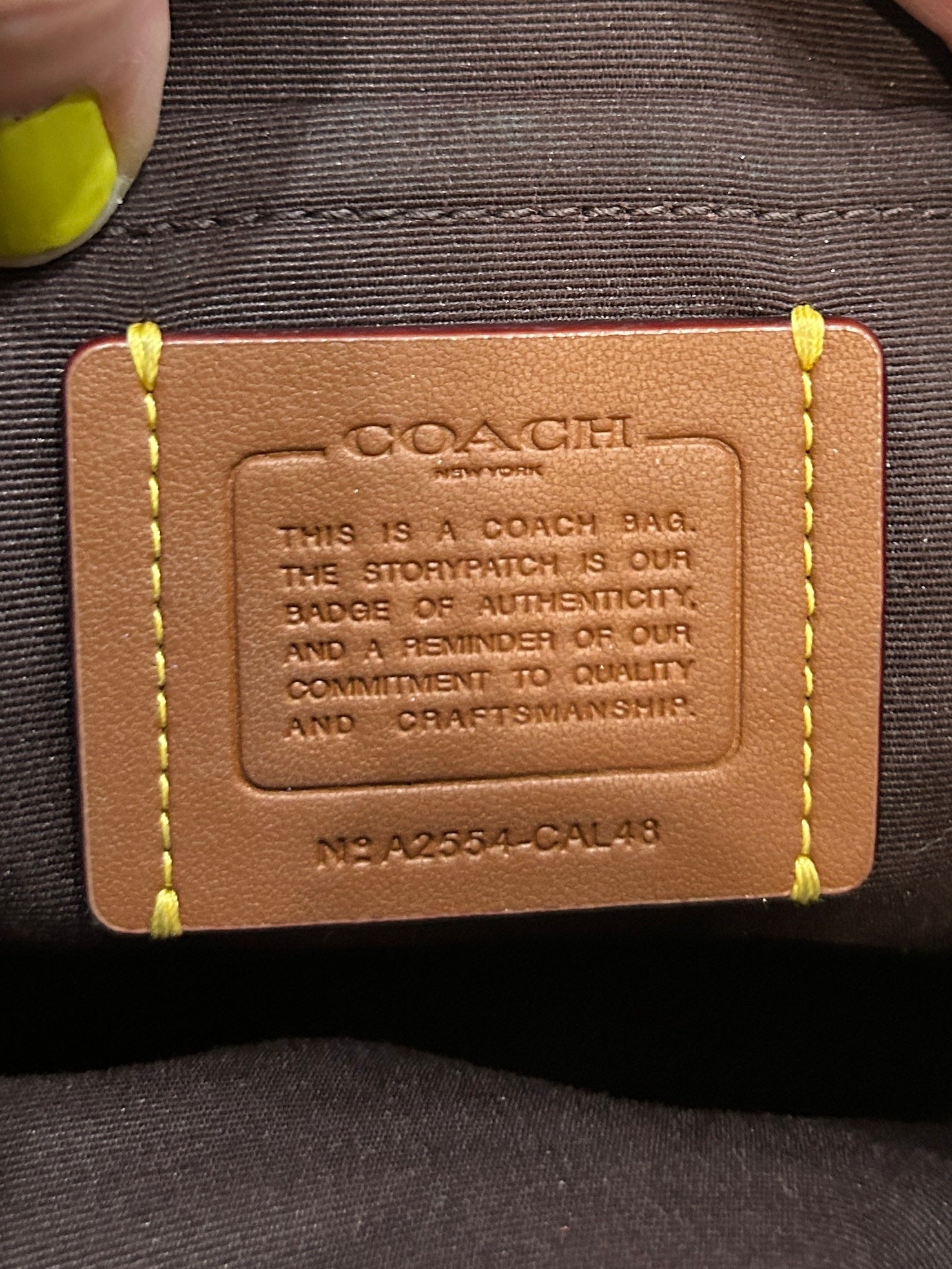 Close-up of a Coach leather authenticity patch sewn onto a dark brown fabric bag, with yellow contrast stitching 