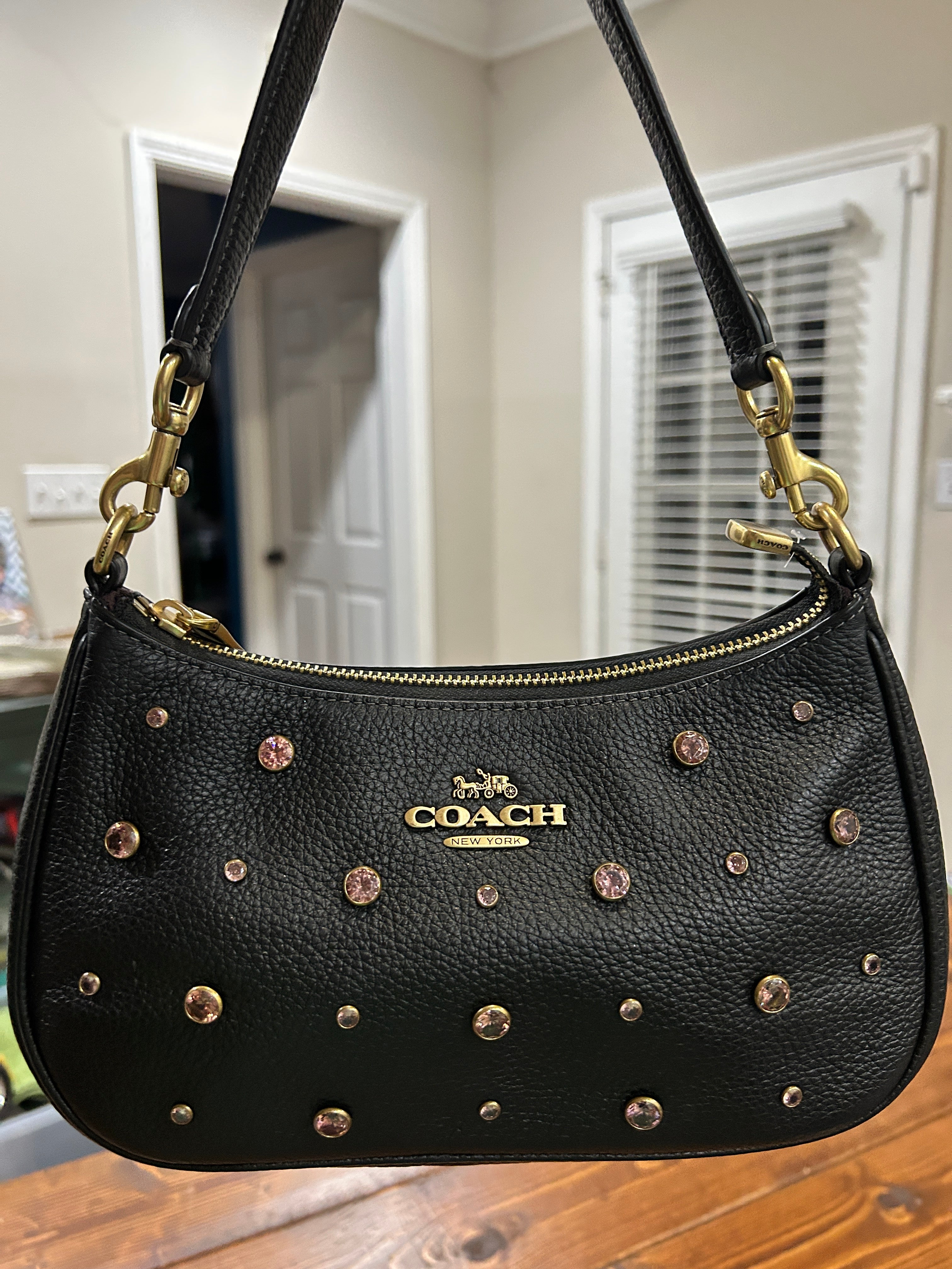 A black pebbled leather Coach shoulder bag with gold-tone hardware and pink faceted crystal embellishments, shown hanging indoors against a home interior background