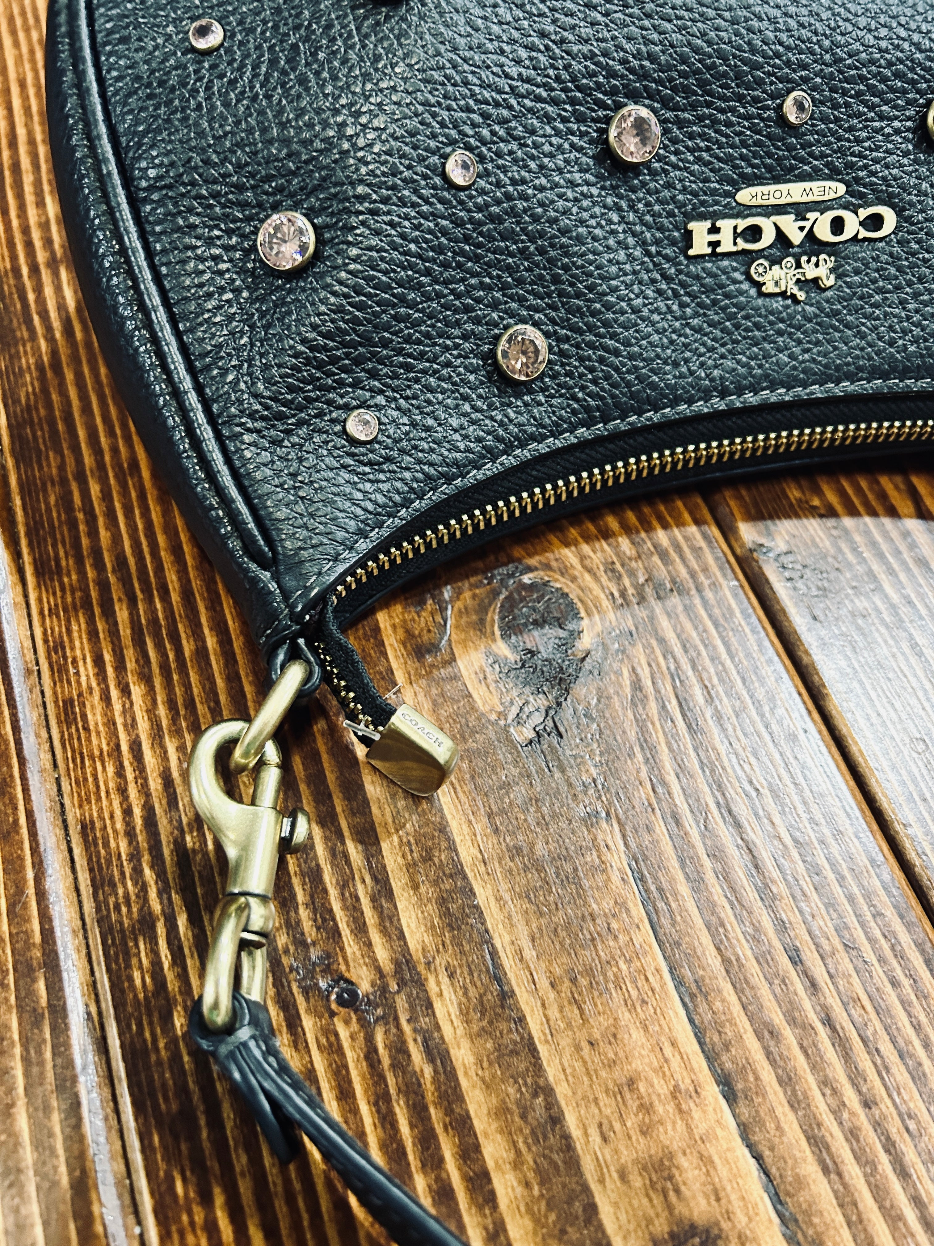Close-up of a black pebbled leather Coach shoulder bag with gold-tone hardware, studs, and a visible Coach logo, resting on a wooden surface.