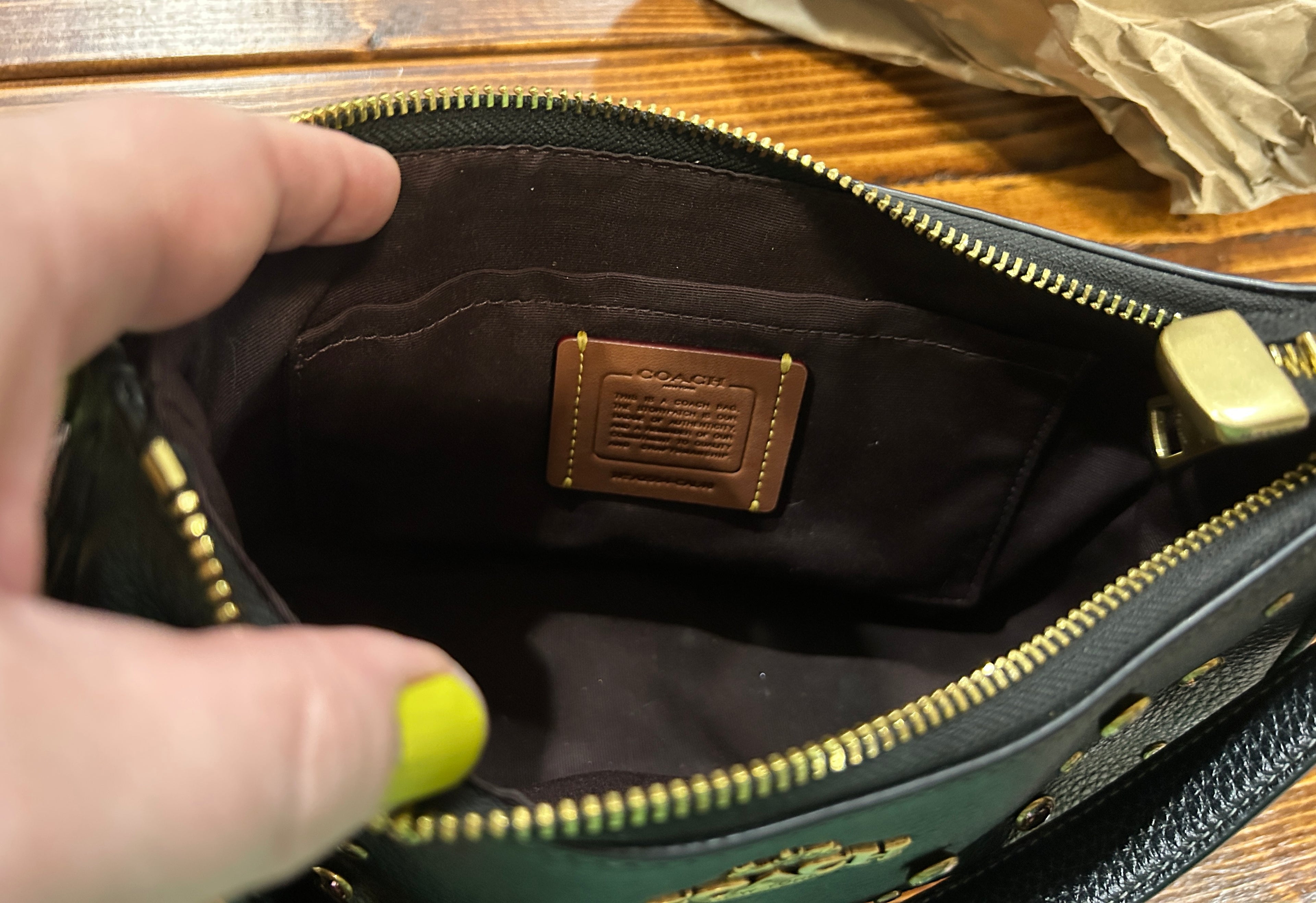 Close-up of an open Coach pouch showing the dark brown interior lining and a branded interior label, with a hand holding the edge and gold-tone zipper hardware visible.