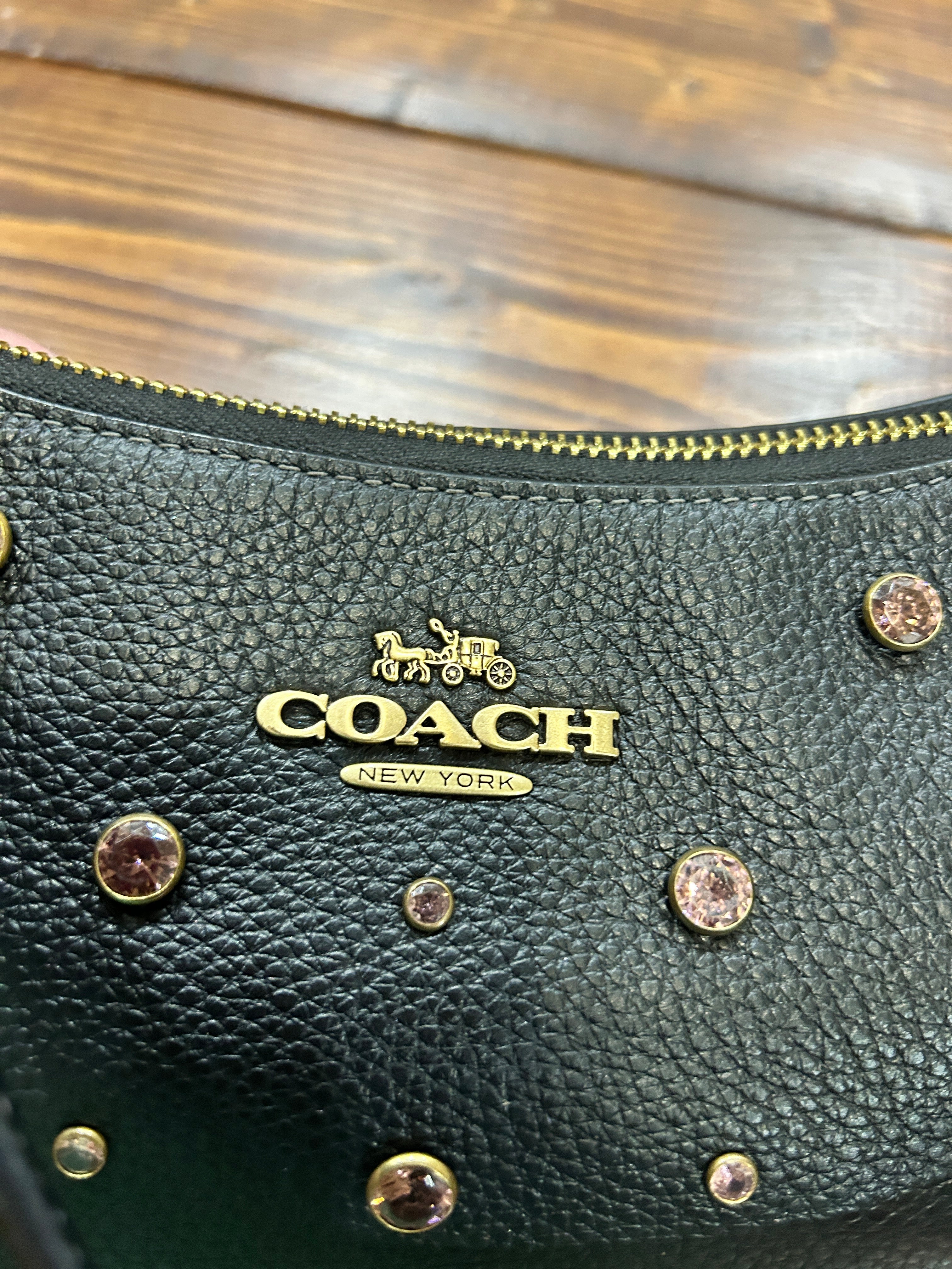 Close-up of a Coach New York handbag in black pebbled leather with gold-tone hardware, including the signature Coach logo, zipper, and decorative crystal studs, resting on a wooden surface.