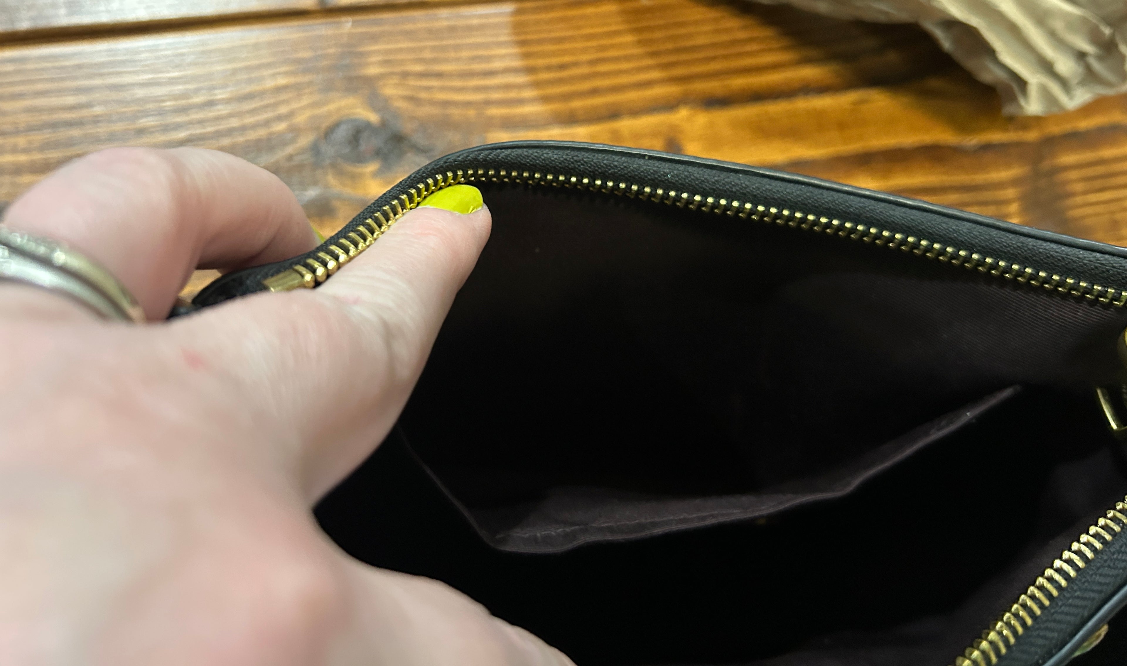 A close-up of a black zippered pouch or clutch being opened by a hand, showing a black interior lining and gold-tone zipper teeth, set against a wooden tabletop background.