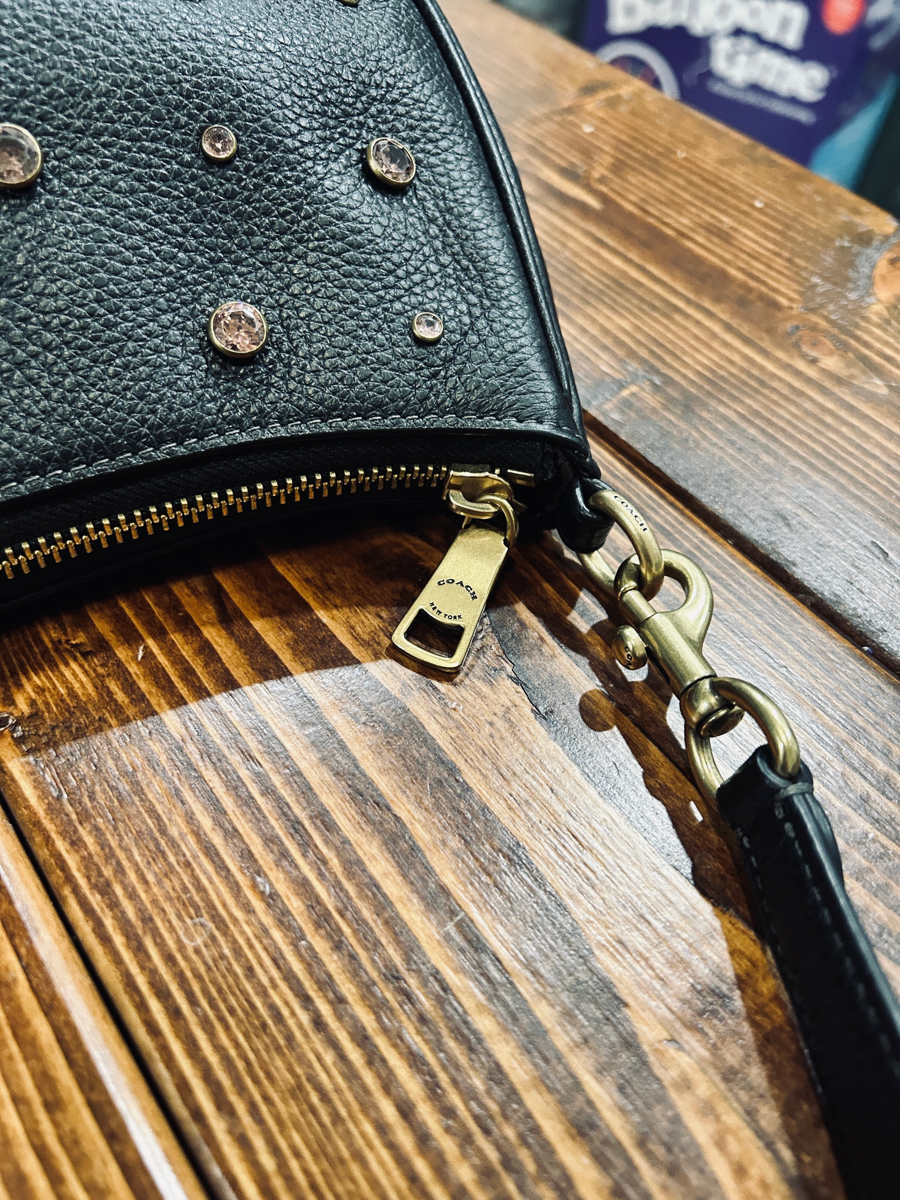 A close-up of a black pebbled leather handbag with gold-tone hardware, including a zipper pull, swivel clasp, and decorative pink crystal studs, resting on a rustic wooden table.
