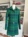 A green and blue patterned wool coat displayed on a mannequin torso in a home setting, with white shelves and framed art in the background.