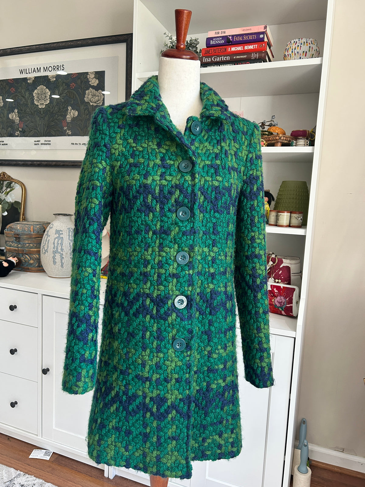 A green and blue patterned wool coat displayed on a mannequin torso in a home setting, with white shelves and framed art in the background.