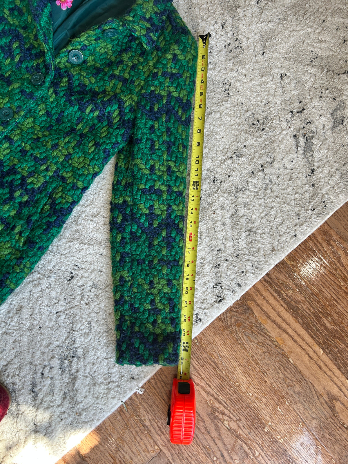 A green and blue patterned knit jacket laid flat on a light rug next to a yellow tape measure, with a wooden floor visible in the corner.