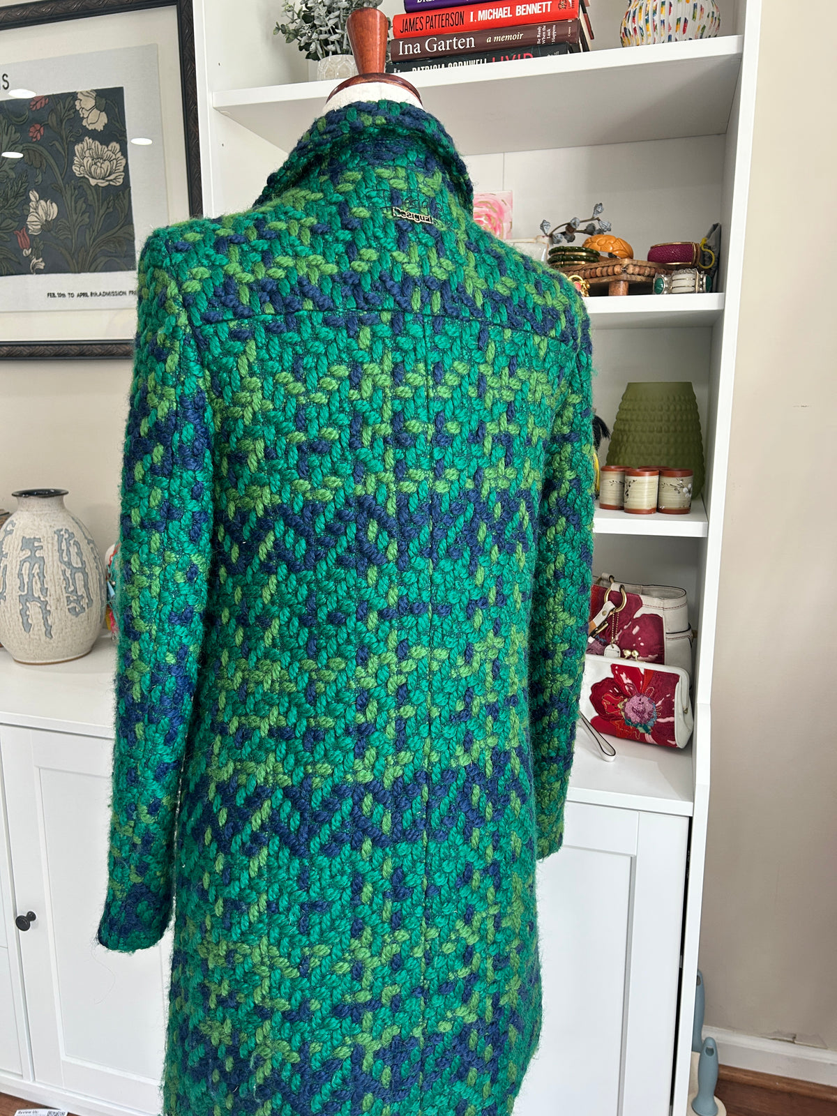A textured, long-line coat with a green and blue knit design displayed on a dress form in front of a white bookshelf.